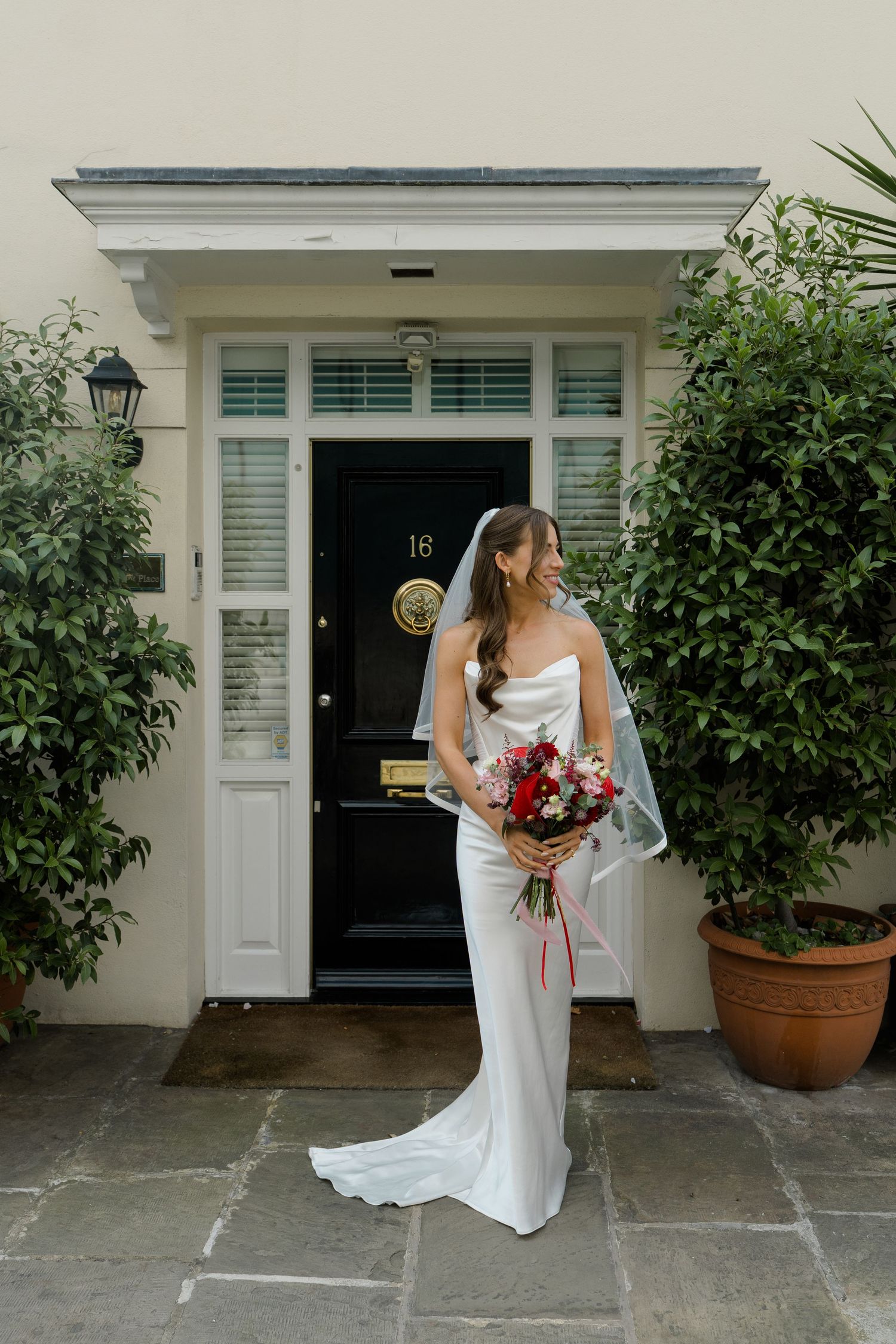 Bride in strapless white gown holding red bouquet stands before black door number 18 with lush greenery.