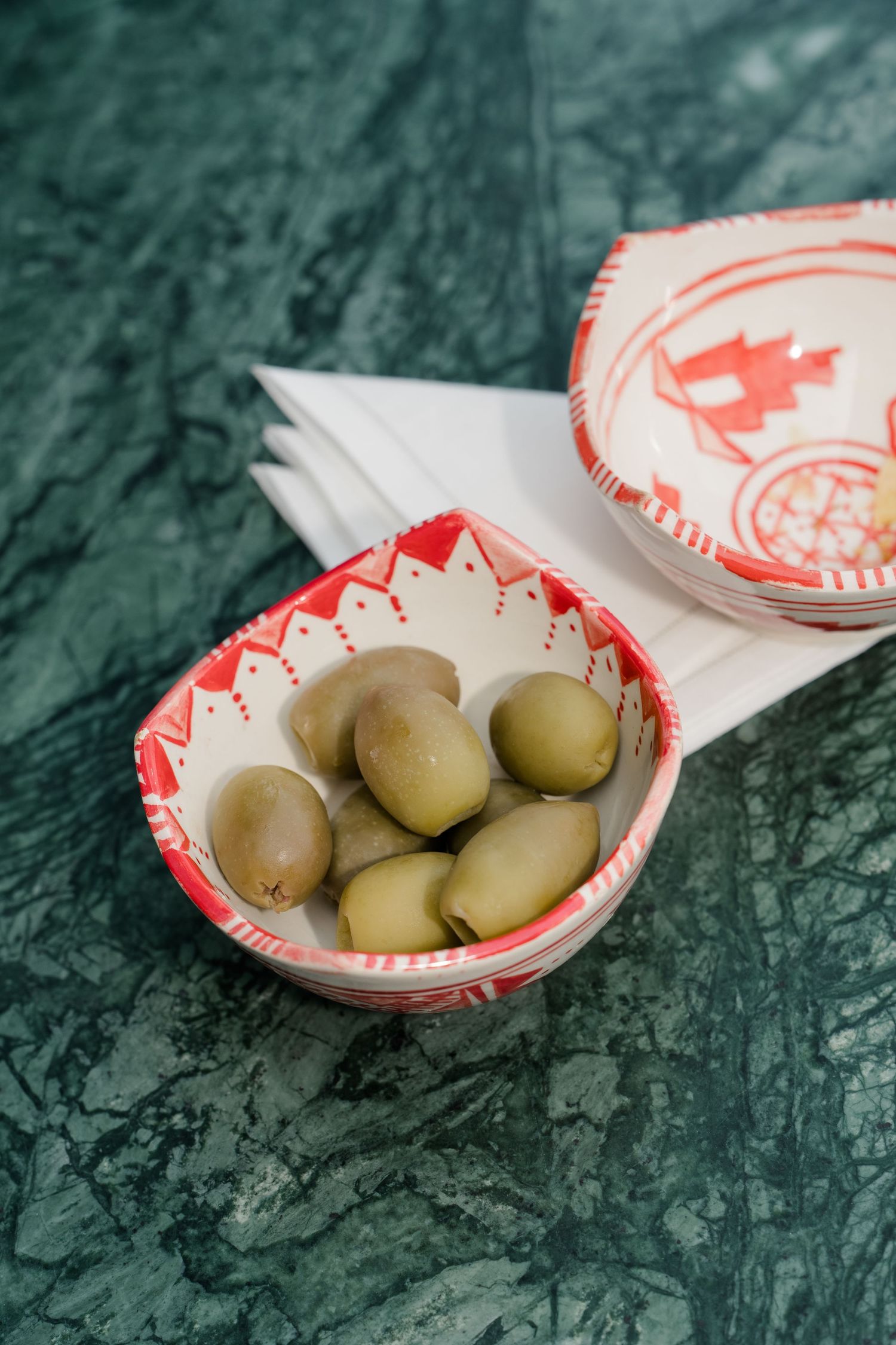 Green olives served in a decorative red and white ceramic bowl on green marble surface.