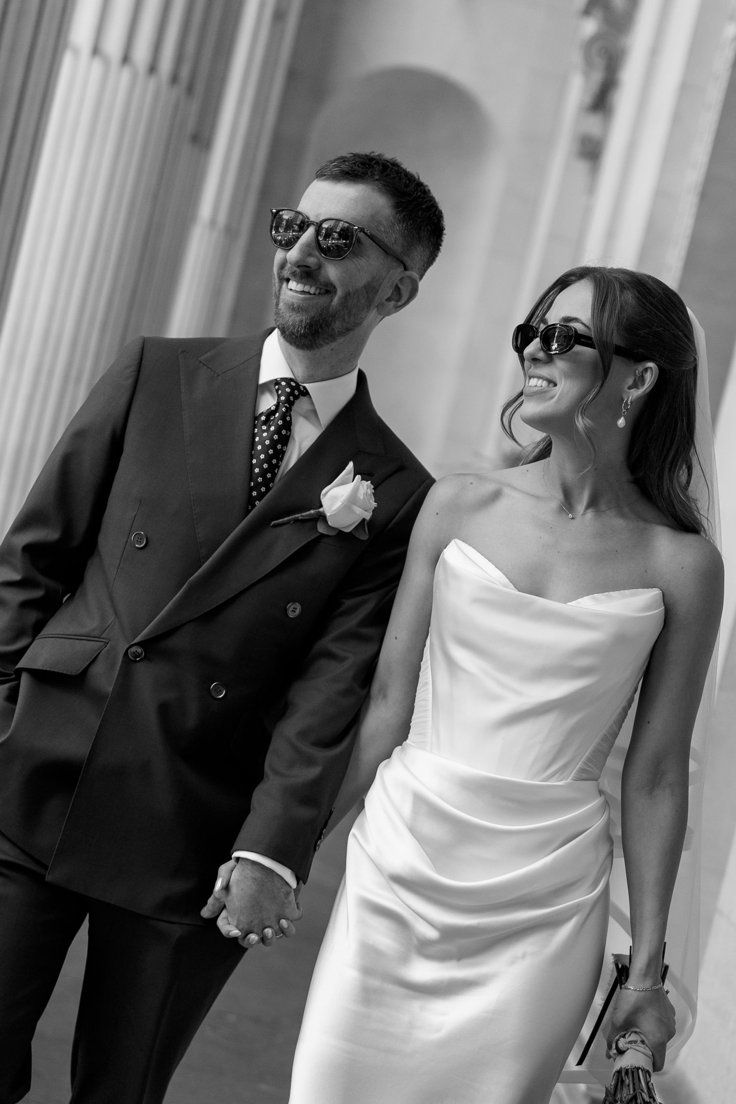 Black and white photo of laughing bride and groom in sunglasses walking between stone columns.