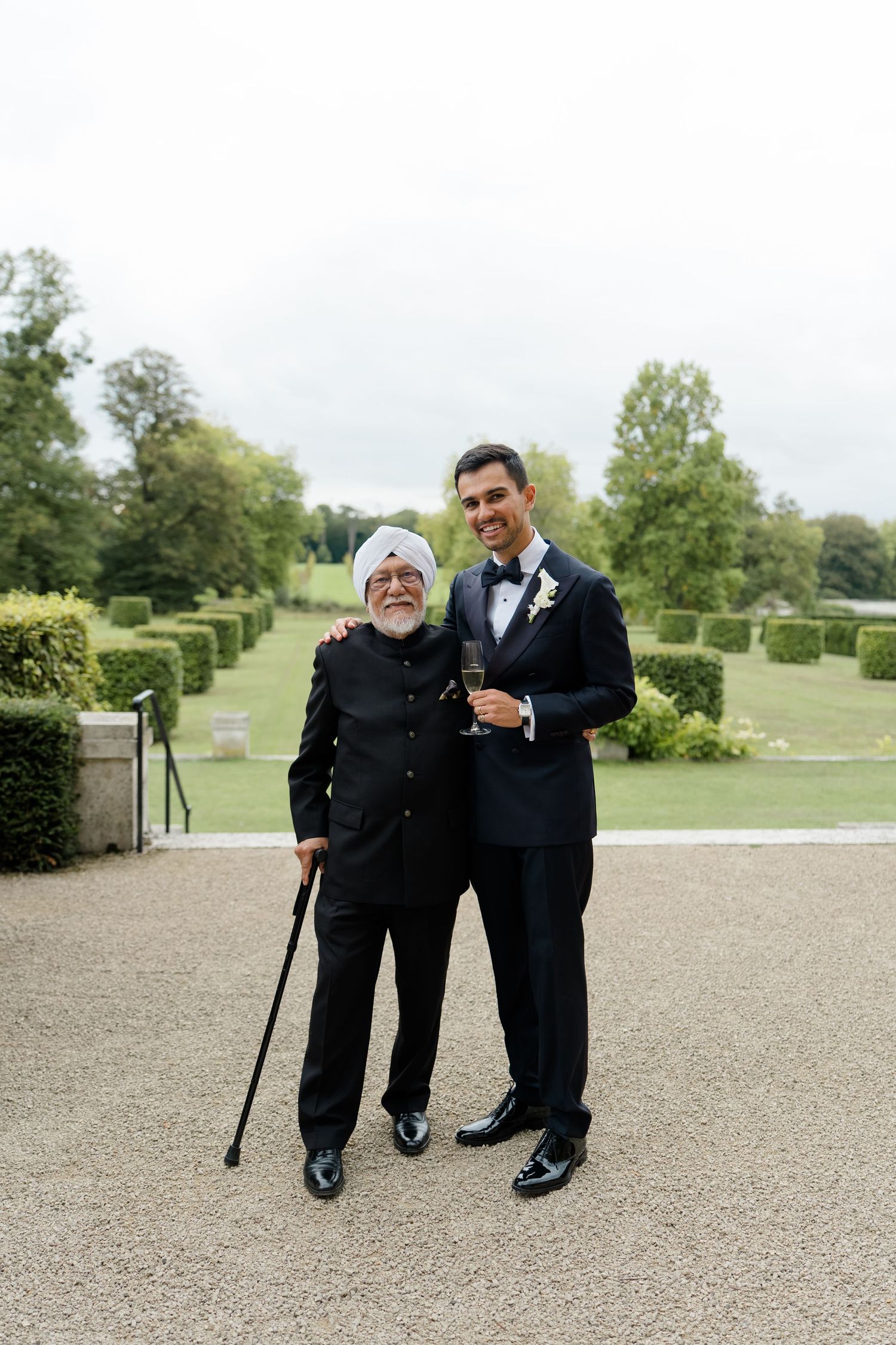 Groom in black tuxedo poses with elderly man in turban and cane in formal garden.