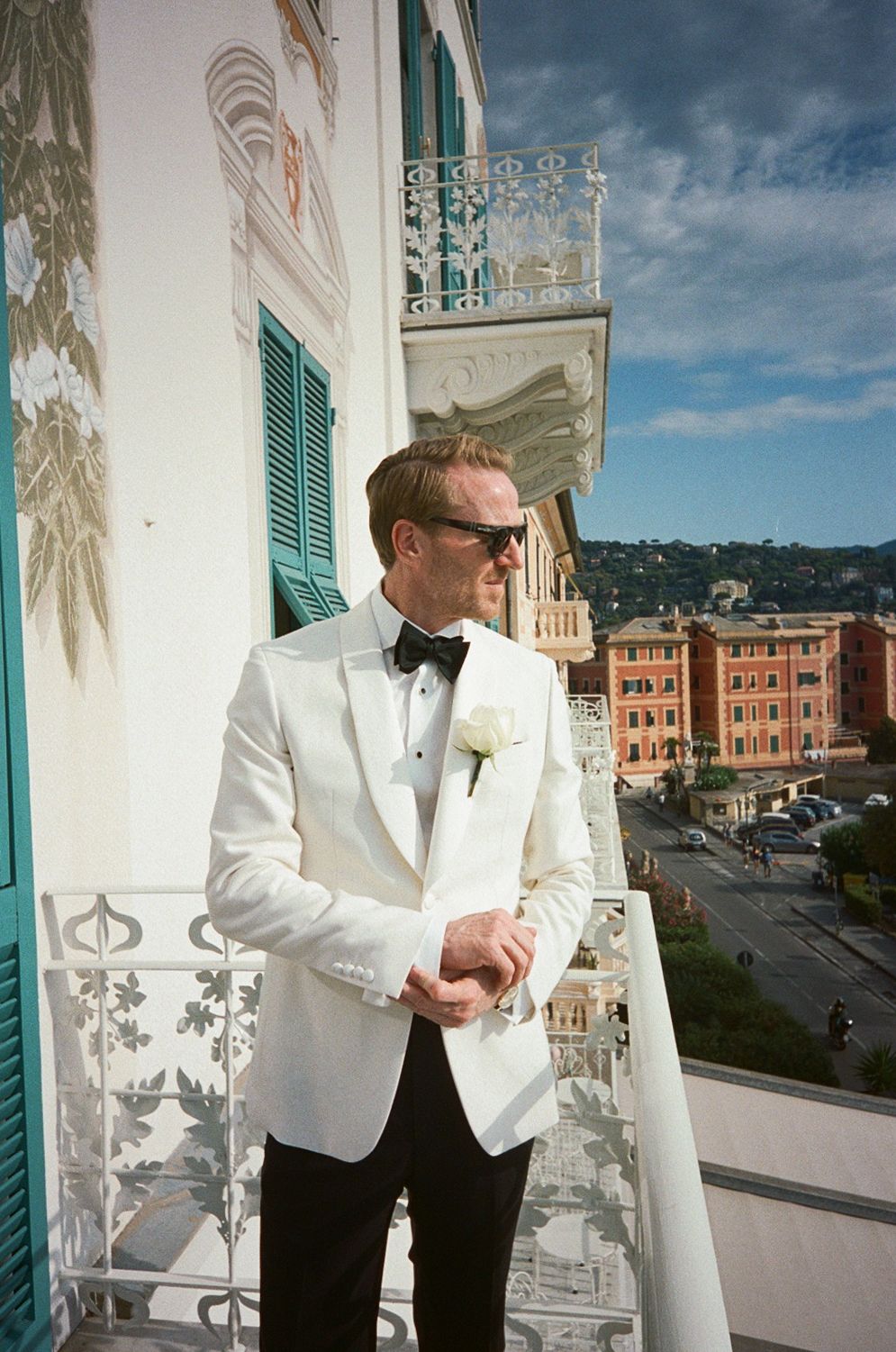 Elegant groom in white tuxedo jacket with boutonniere stands on hotel balcony overlooking coastal town.
