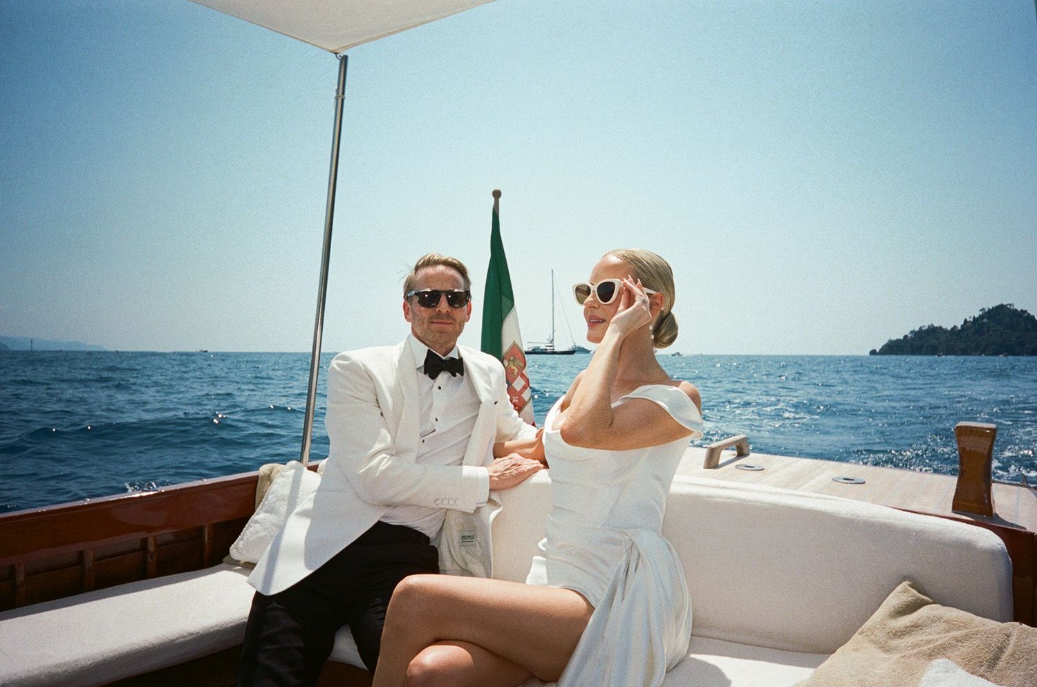Elegant bride and groom in white wedding attire relax on a luxury boat on the Italian Riviera coastline.