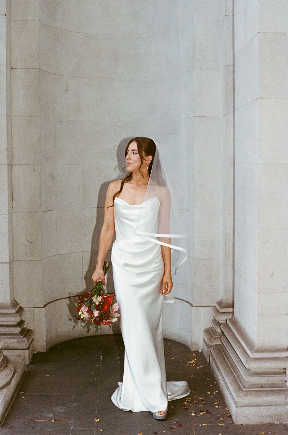 Elegant bride in sleek white gown with veil poses between stone pillars holding floral bouquet.