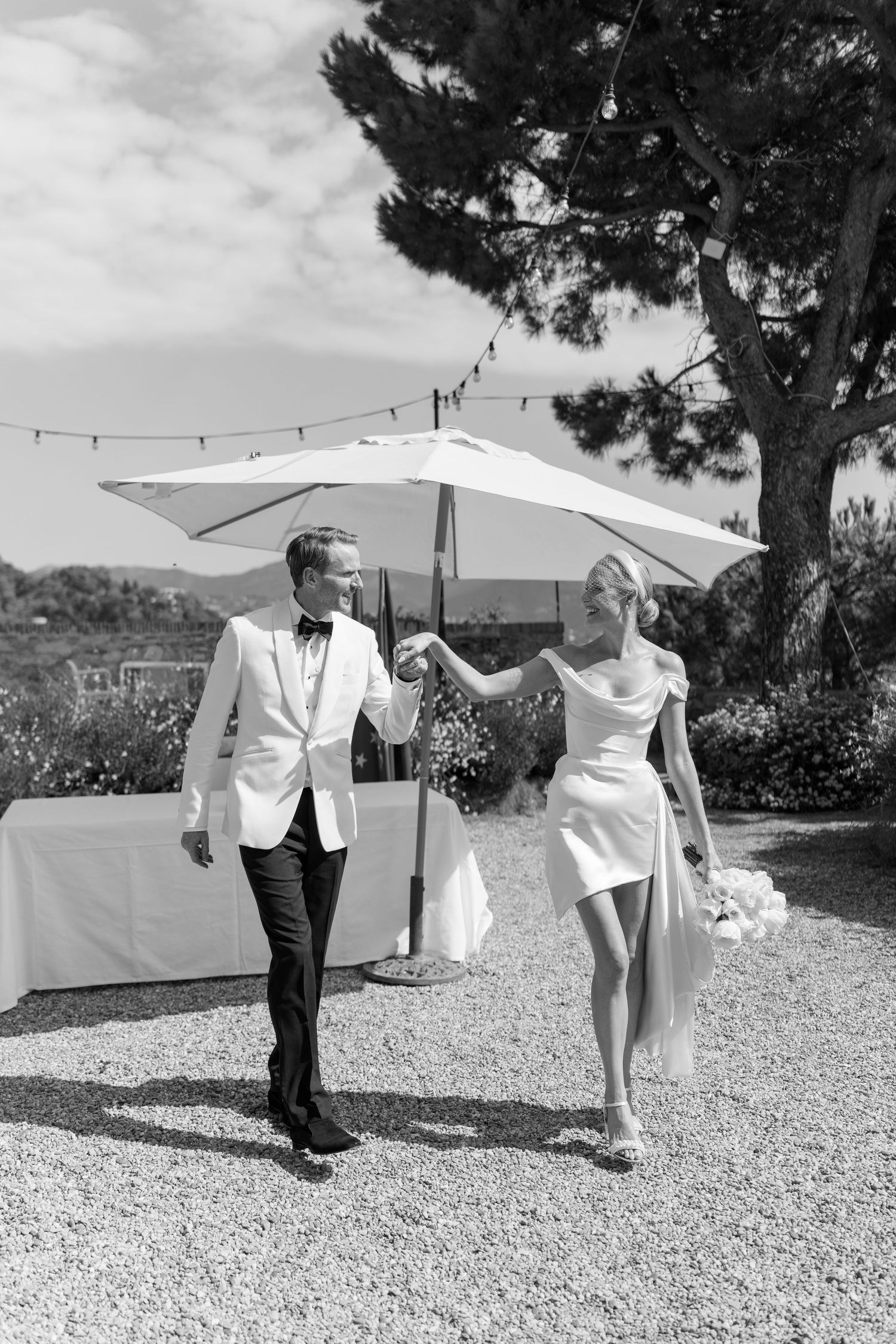 Black and white photo of bride and groom walking hand in hand on gravel path at outdoor venue.