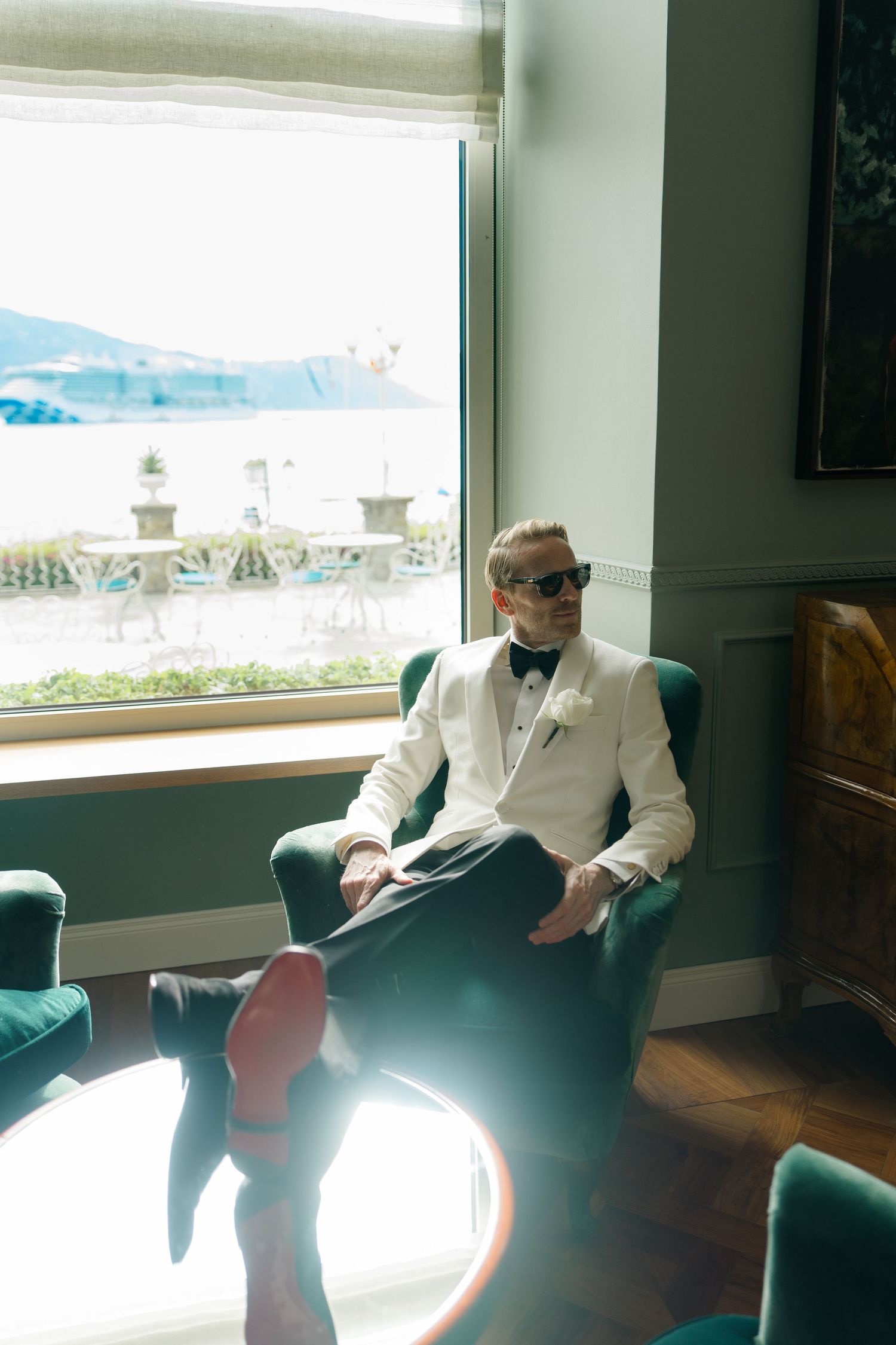 A groom in a white tuxedo jacket and bow tie sits in a green velvet chair by a large sunlit window.