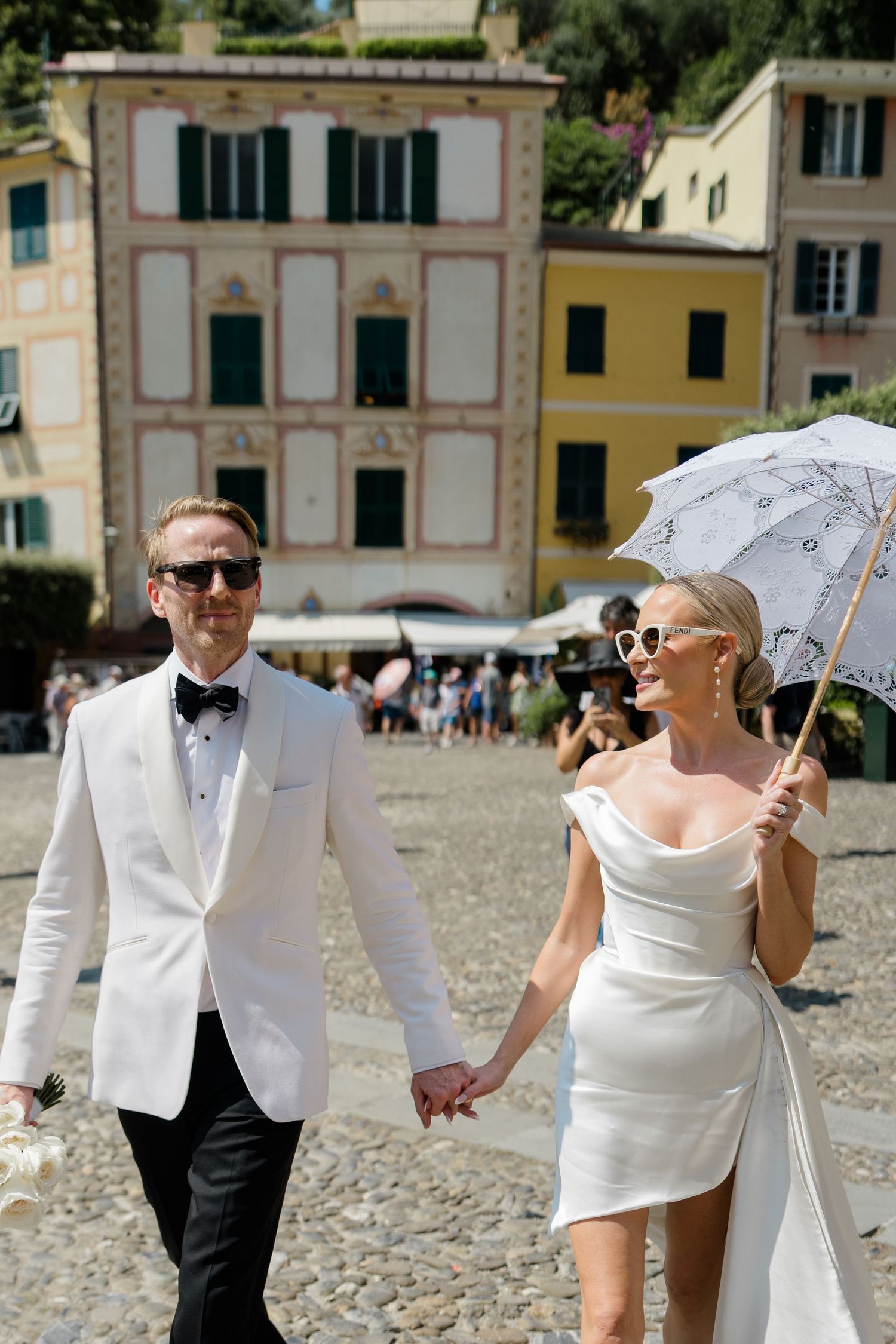 Bride and groom walking hand in hand through colorful Italian piazza, bride holding lace parasol.