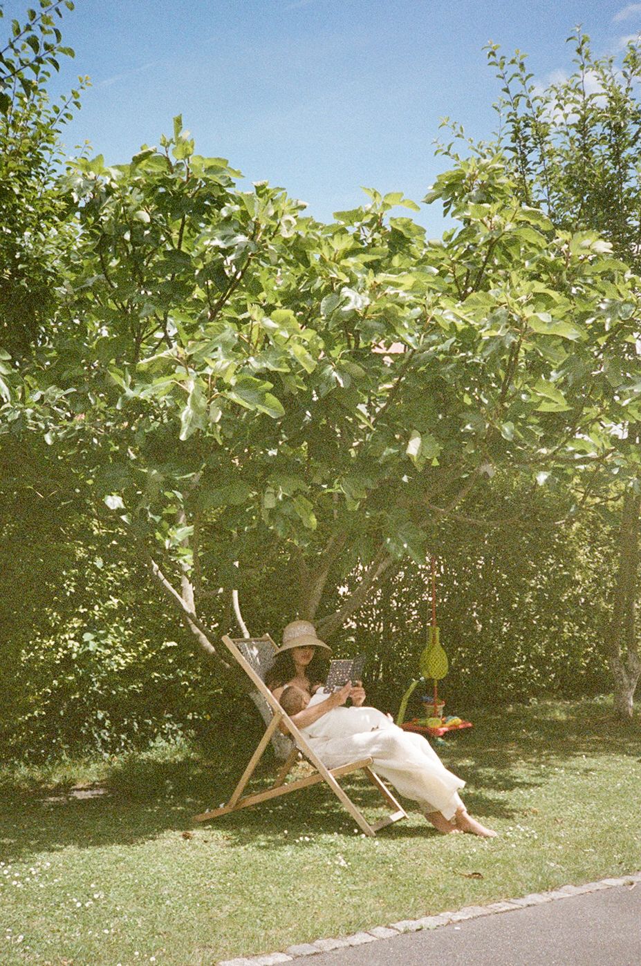 Woman in white dress and hat relaxing on wooden deck chair in sunny garden reading a book.