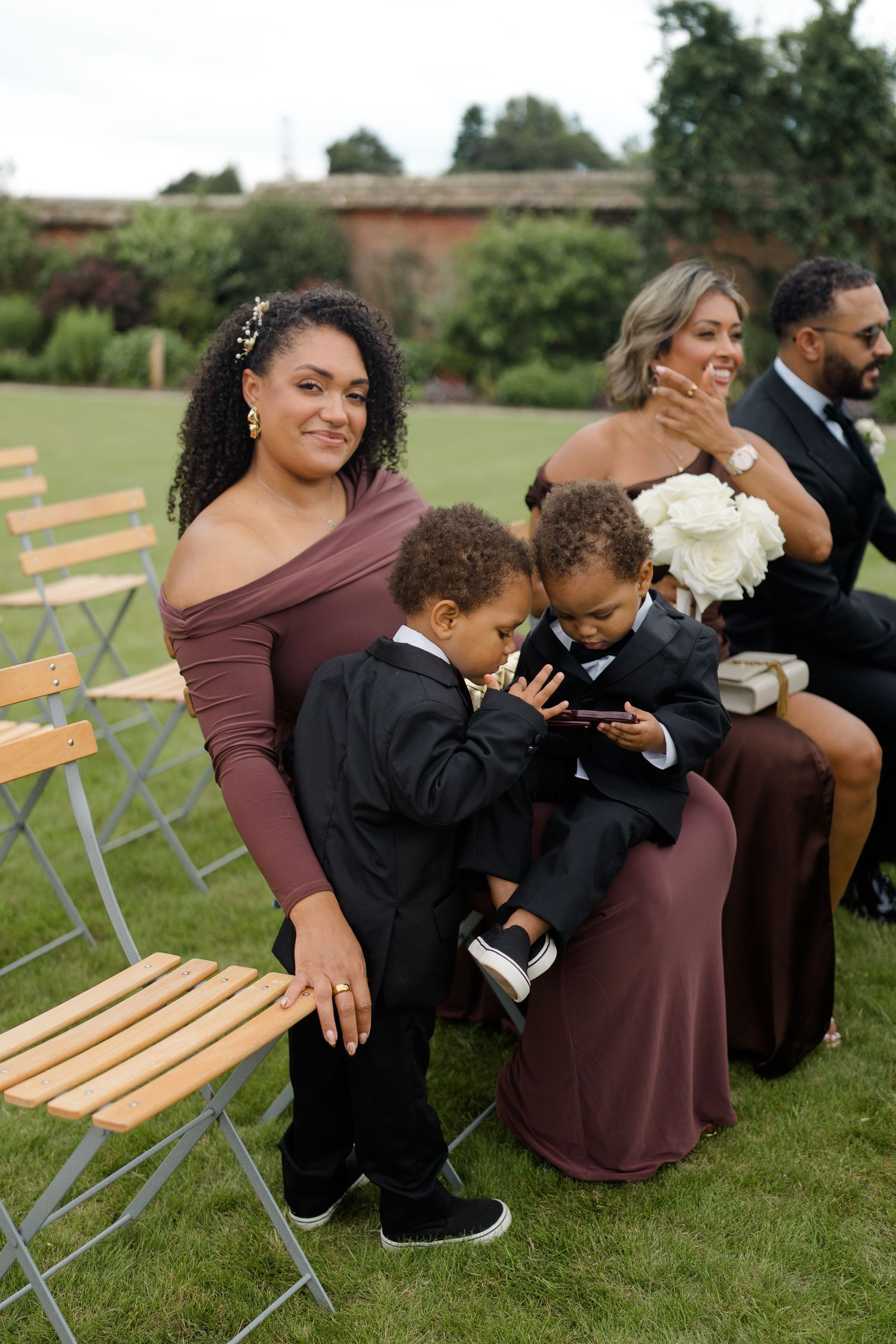 A woman in a brown dress sits with two young boys in suits at an outdoor wedding.