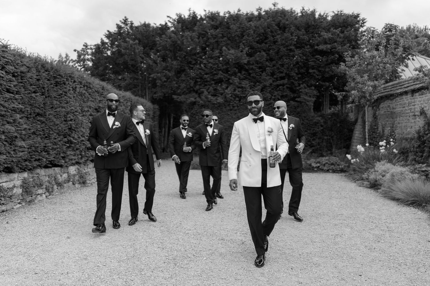 Groom in white jacket leads groomsmen in black tuxedos along garden path, black and white photo.