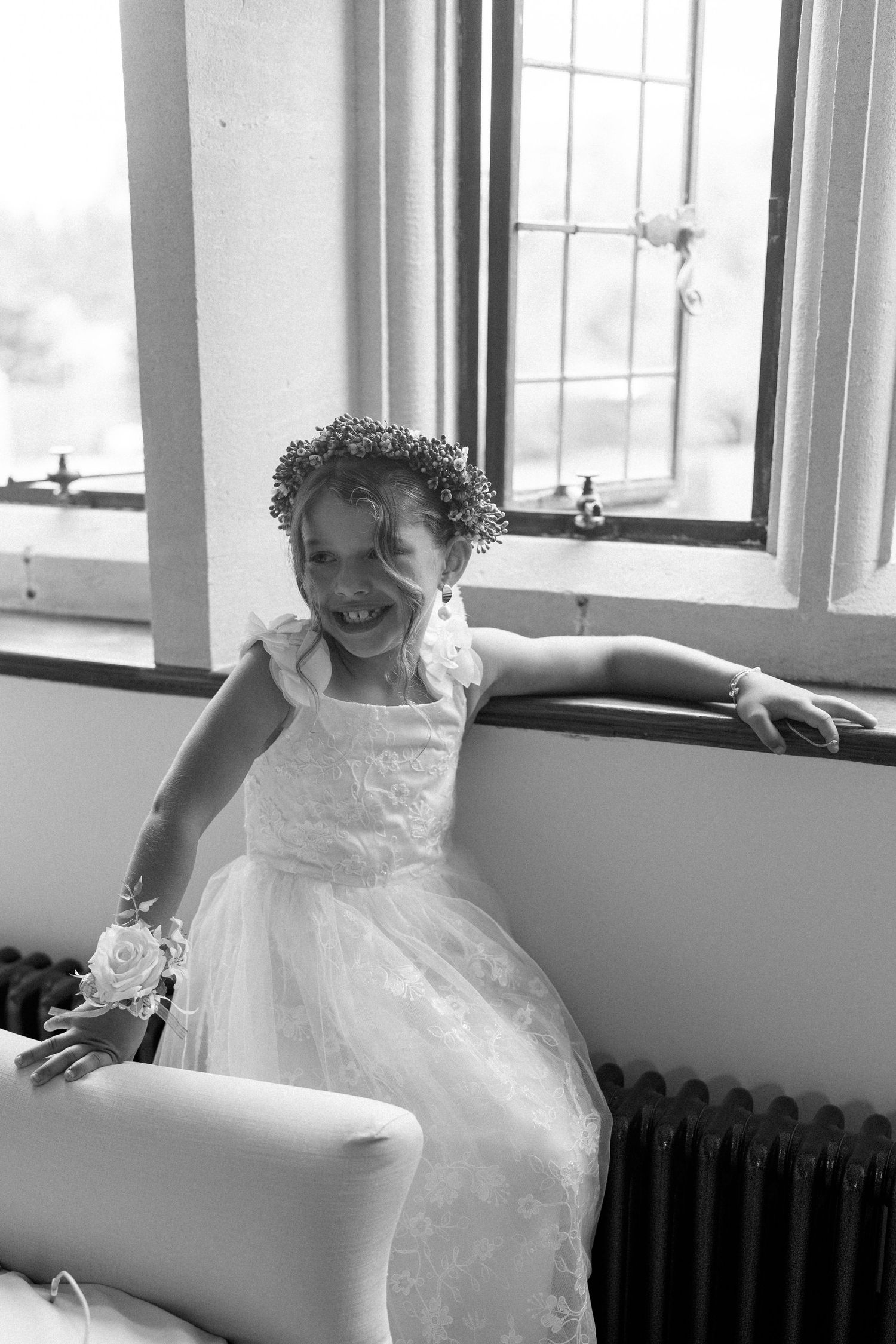 Black and white portrait of a smiling flower girl in a white dress with a floral crown.