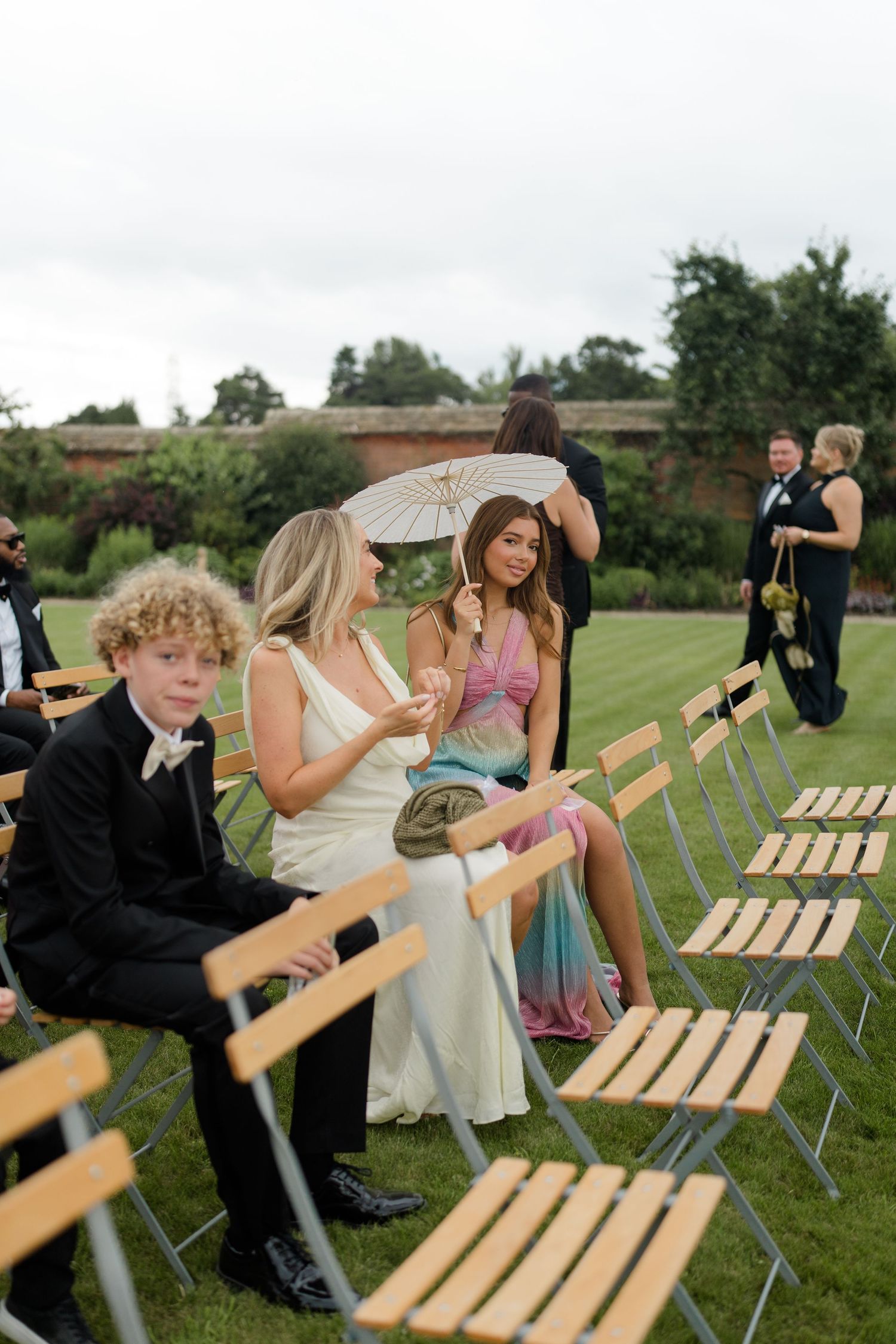Wedding guests seated in wooden folding chairs at outdoor ceremony, woman holding white parasol in colorful dress.