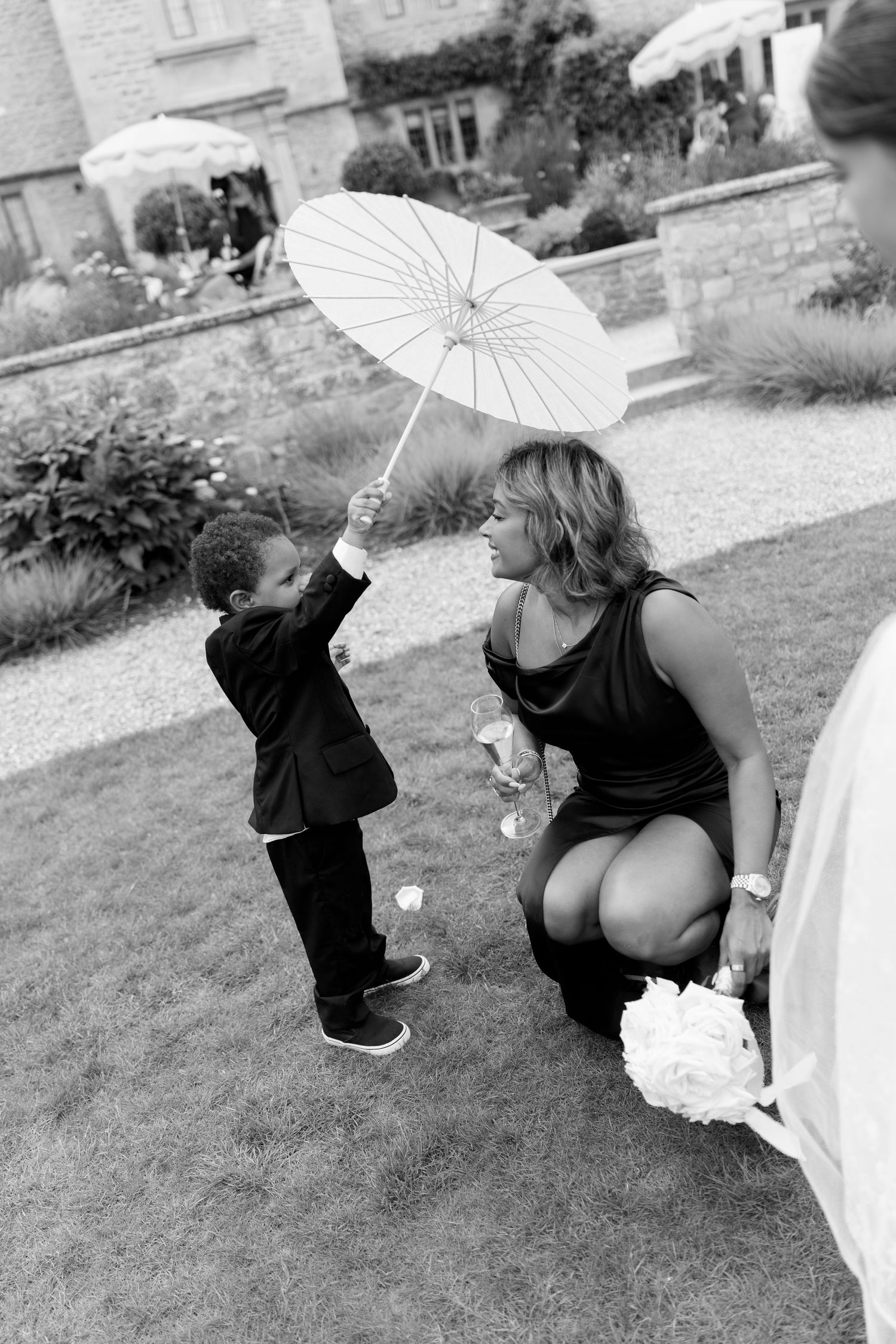 Young boy in suit holds white parasol for bridesmaid crouching down in black and white wedding photo.