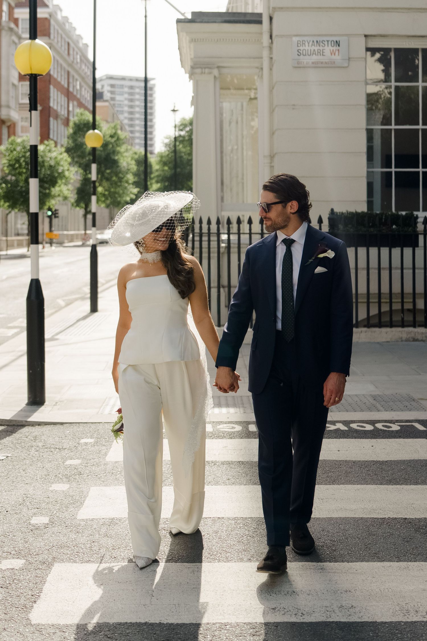 Bride and groom in white suit and navy blue hold hands crossing street at Bryanston Square London.