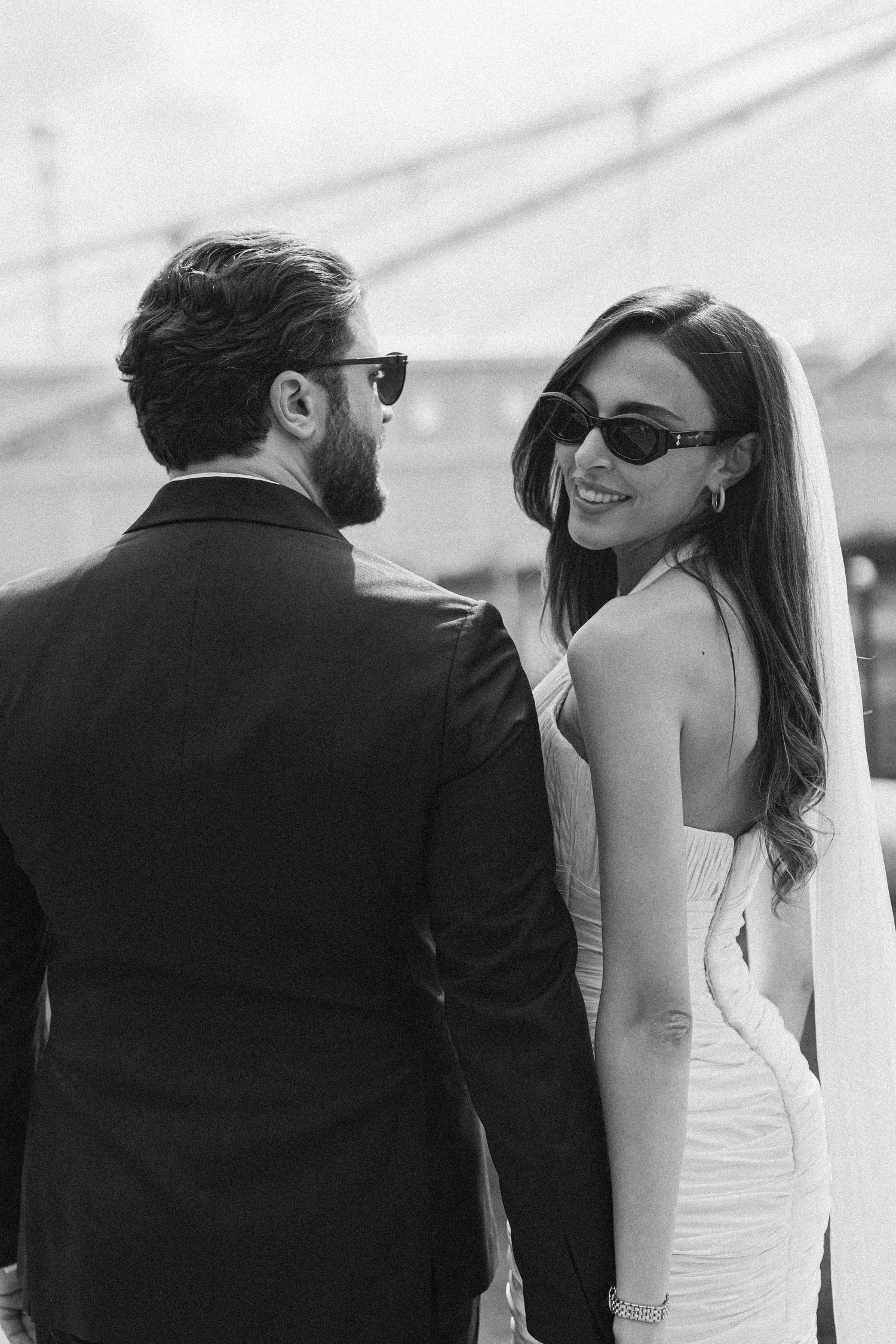 Black and white photo of a smiling bride in sunglasses and veil with groom in suit at wedding.