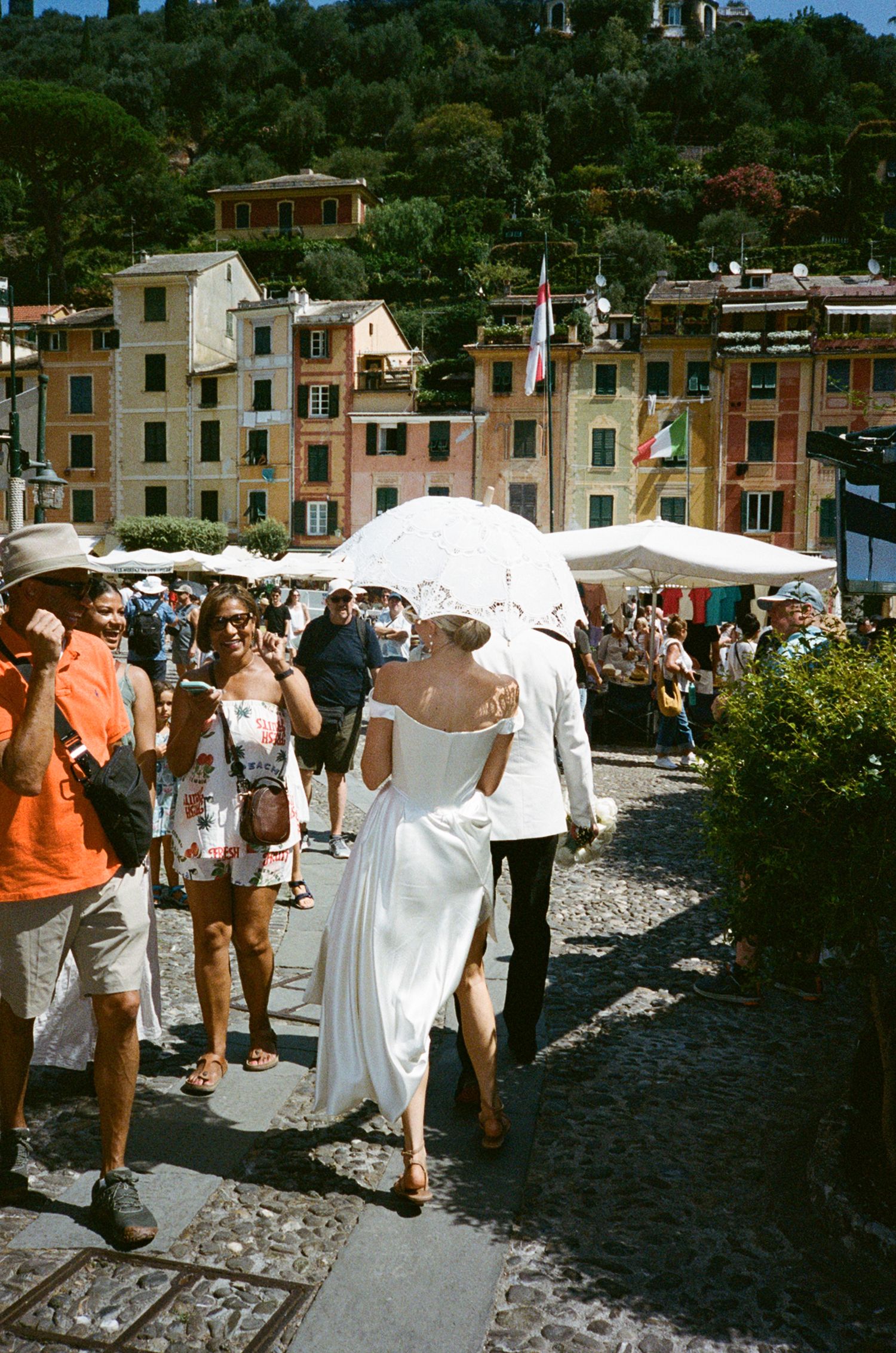 Bride and groom walk under white umbrella in sunny Portofino, Italy, surrounded by colorful buildings and crowds.