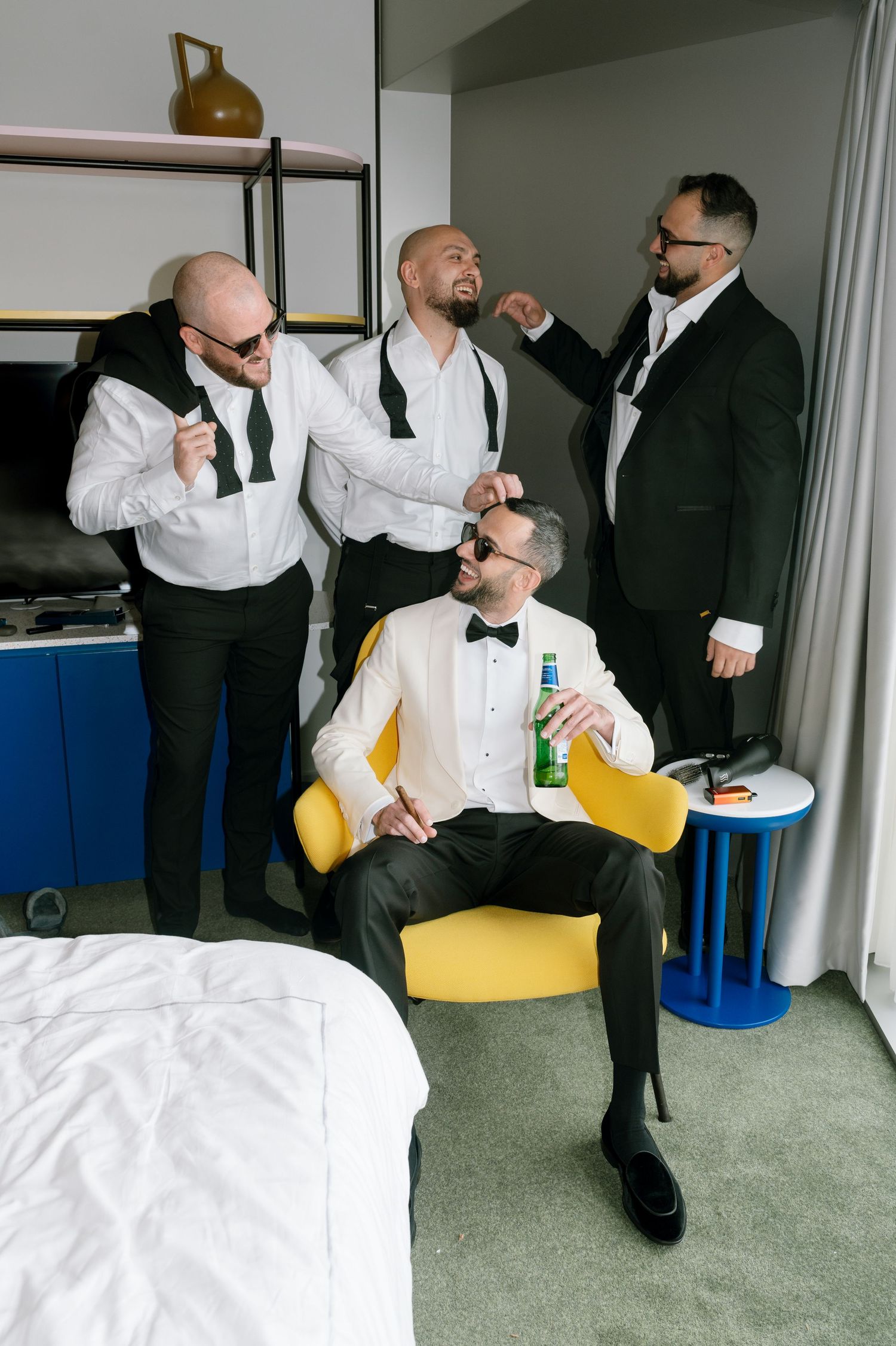 Groomsmen in formal attire getting ready in a hotel room with a yellow chair and blue cooler.
