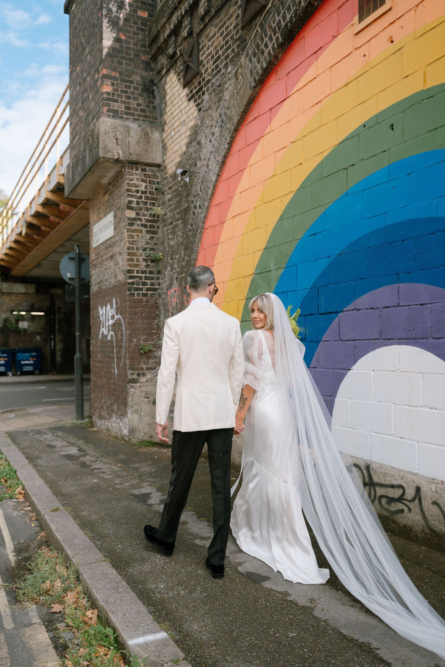 A couple poses against a vibrant rainbow mural on a brick wall, with the bride wearing a flowing white gown and veil.