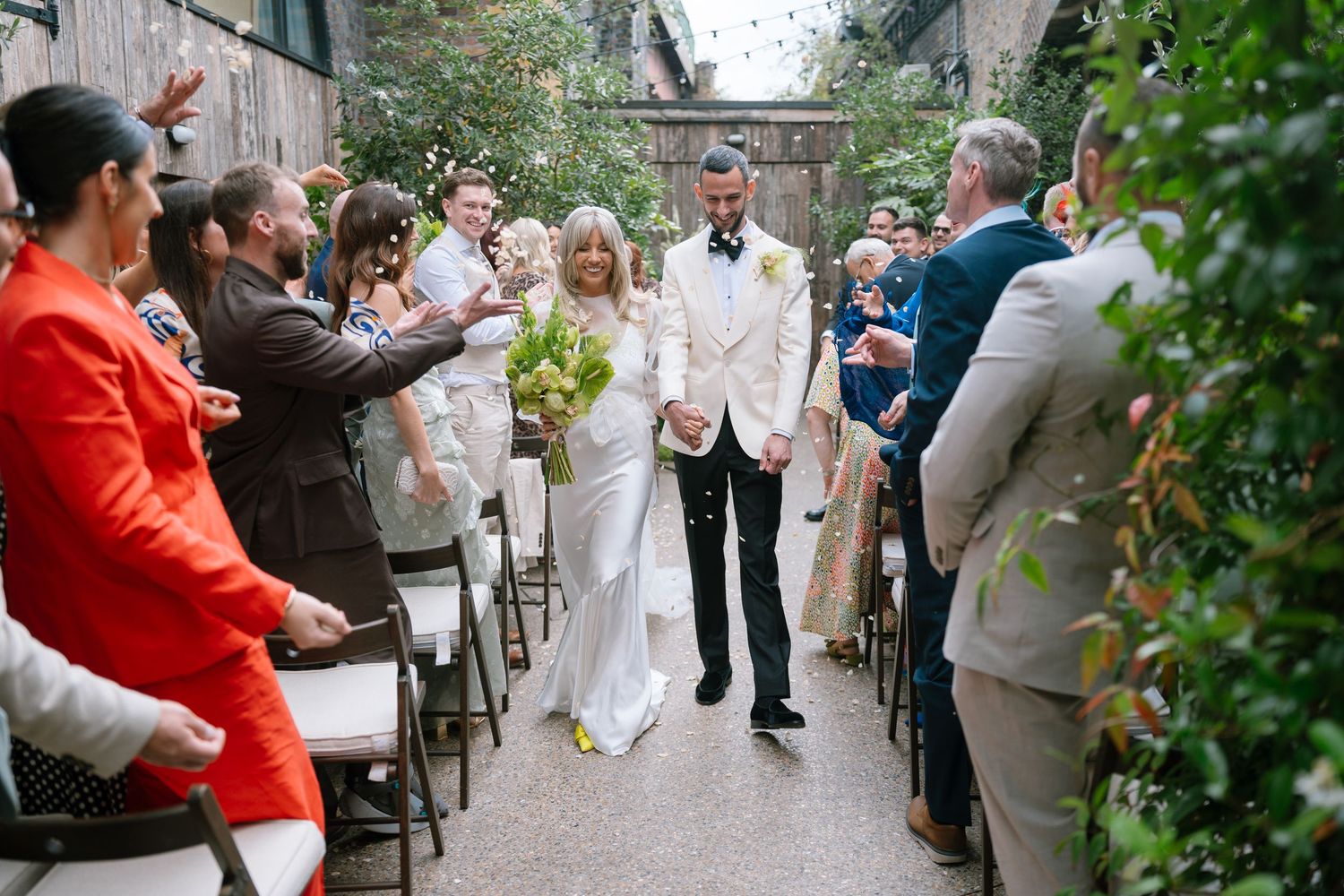 Wedding guests gather outdoors in a garden setting with someone in red standing out in the crowd.