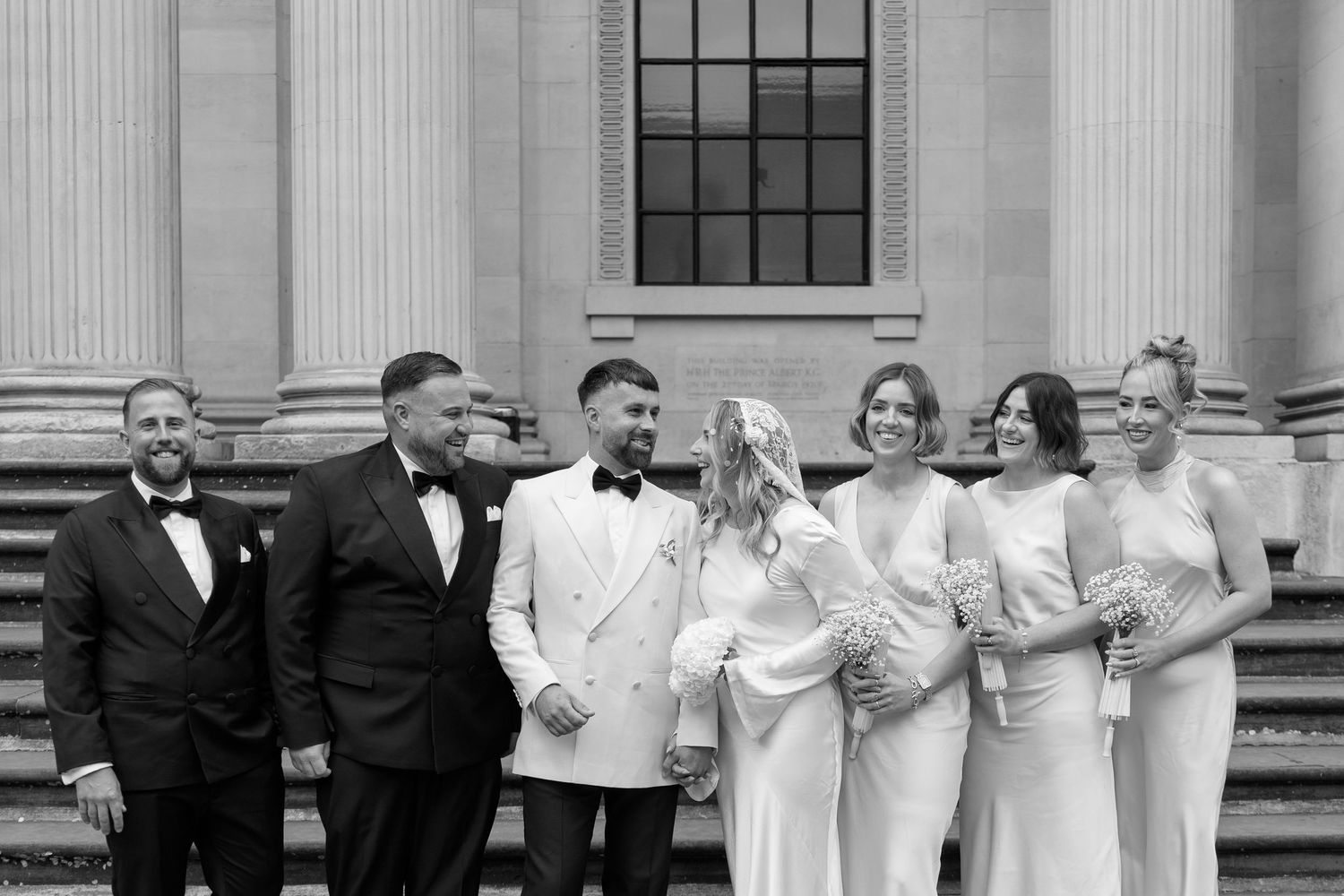 Wedding party in formal attire poses together on steps of classical building with white columns.