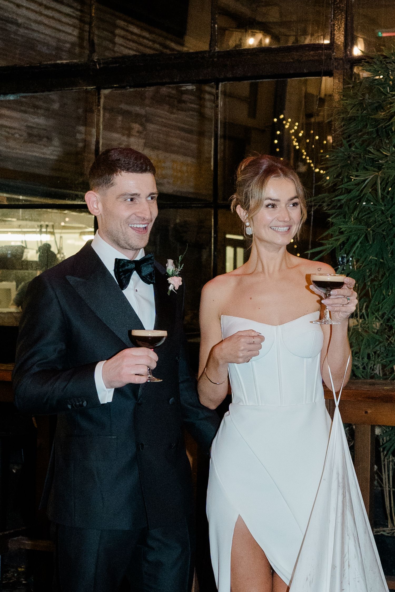 A couple in wedding attire share a toast with wine glasses at an indoor reception venue with twinkling lights.