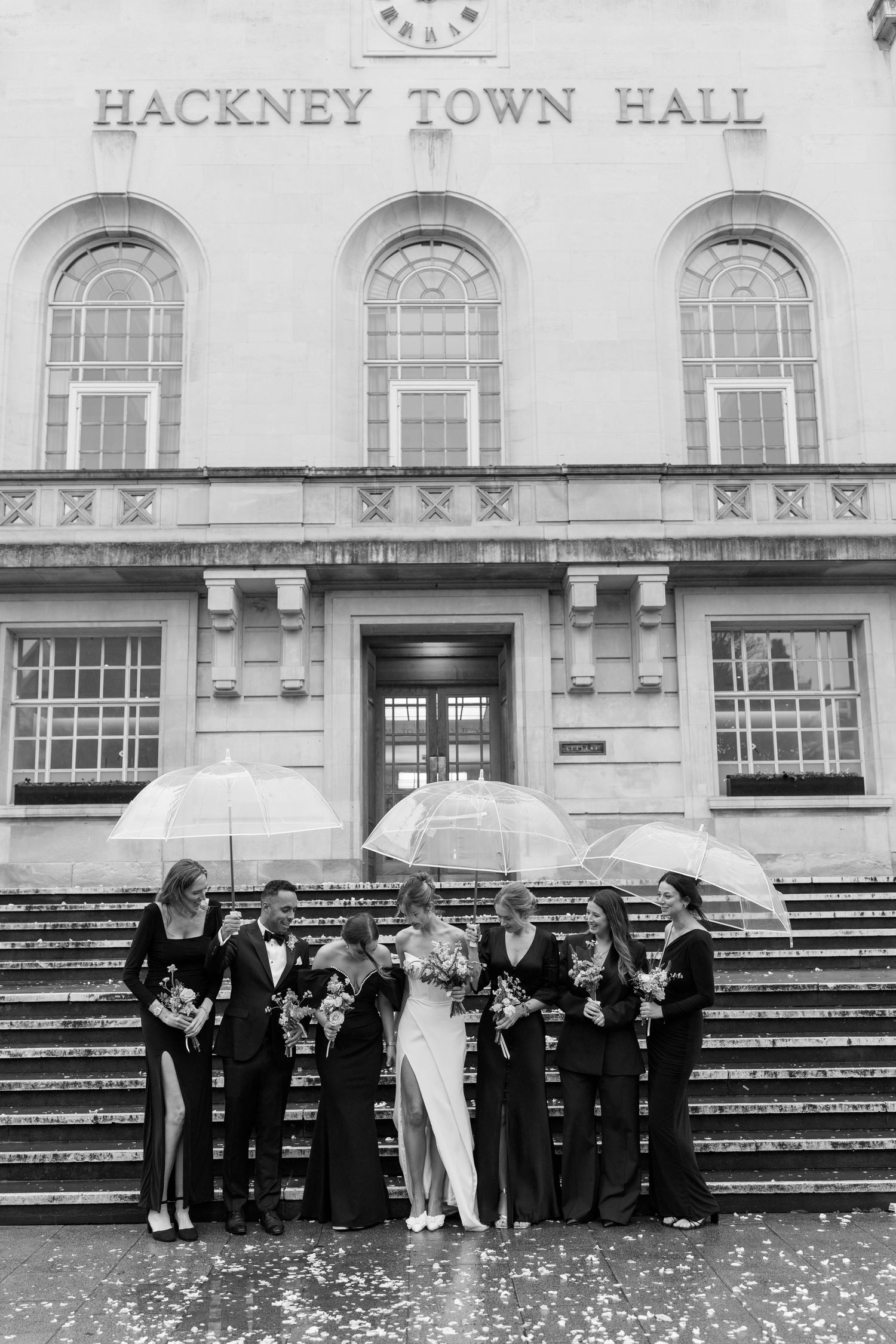A wedding party stands with umbrellas on the steps of Hackney Town Hall in black and white photography.