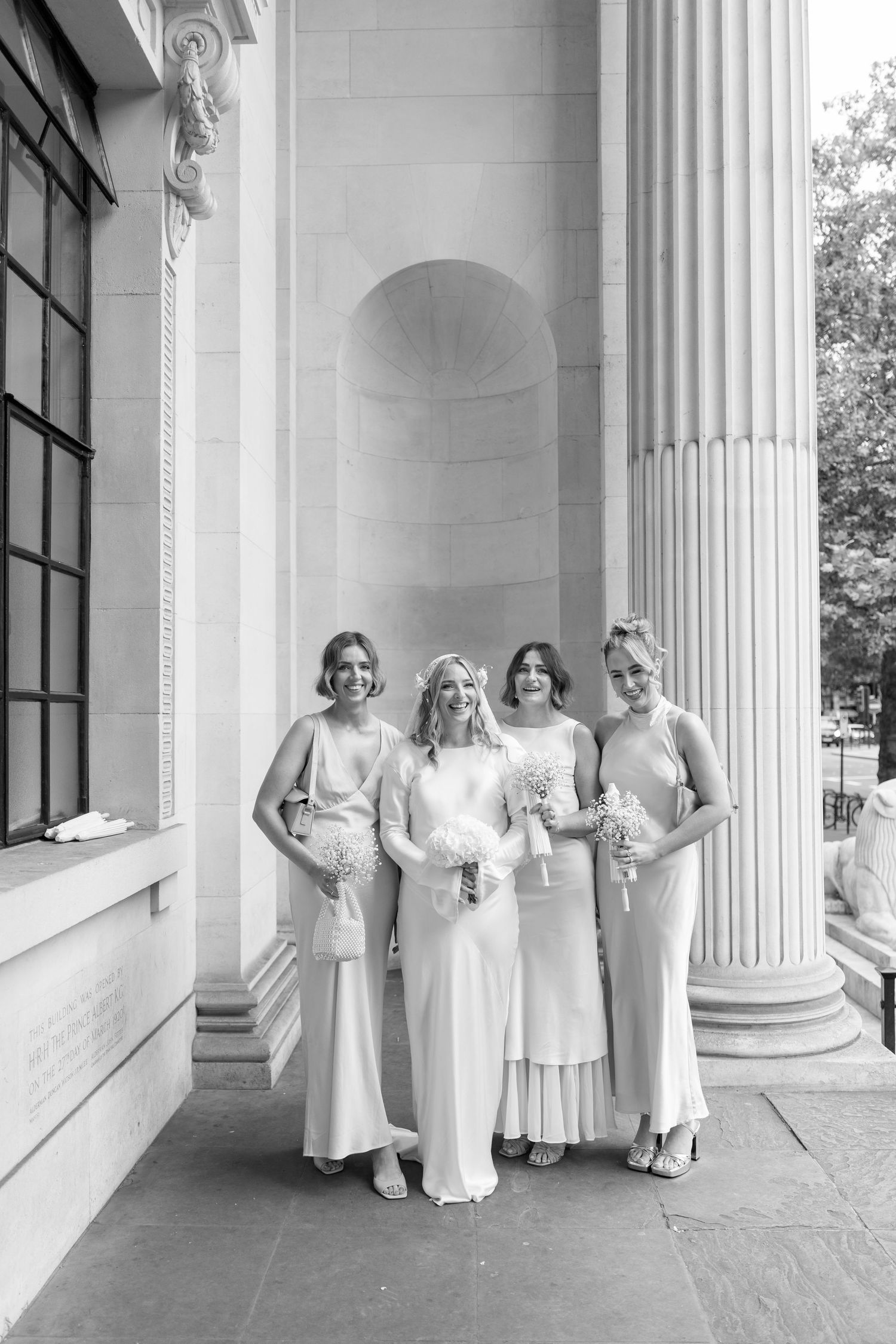 Four bridesmaids in elegant white dresses pose together in front of classical columns at a formal wedding venue.