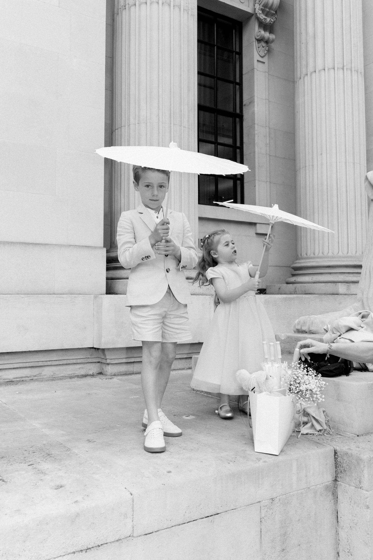 Two children in white formal attire hold parasols while standing on stone steps near white columns.