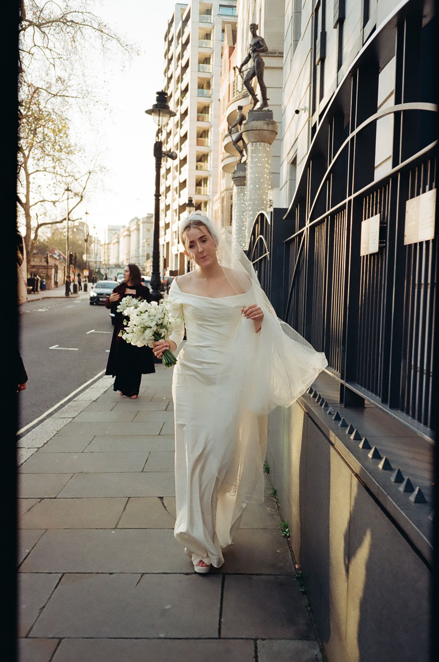 A person in a white wedding dress walking along an urban sidewalk with tall buildings.