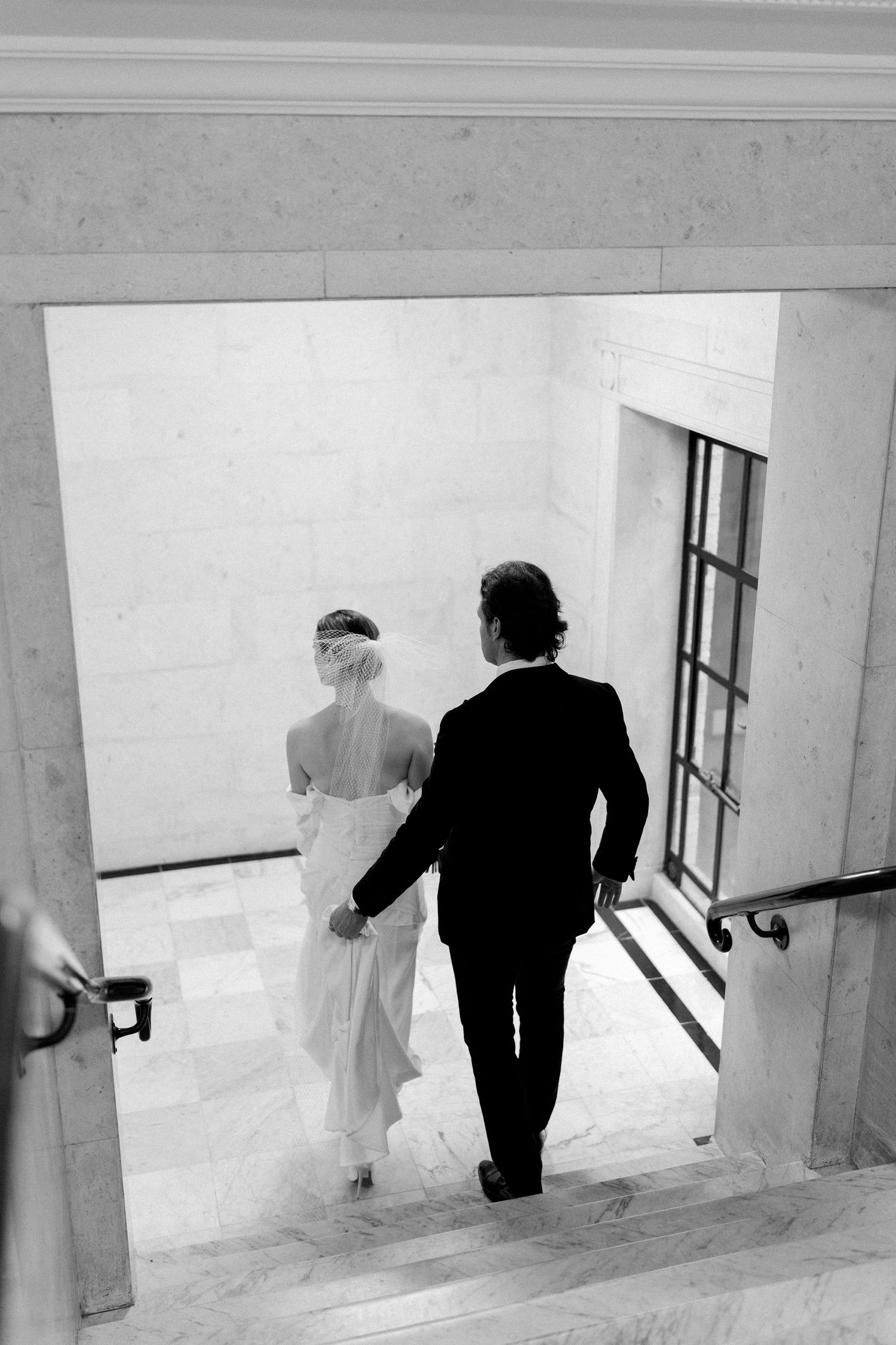 A black and white photograph of a couple descending marble stairs in formal attire.