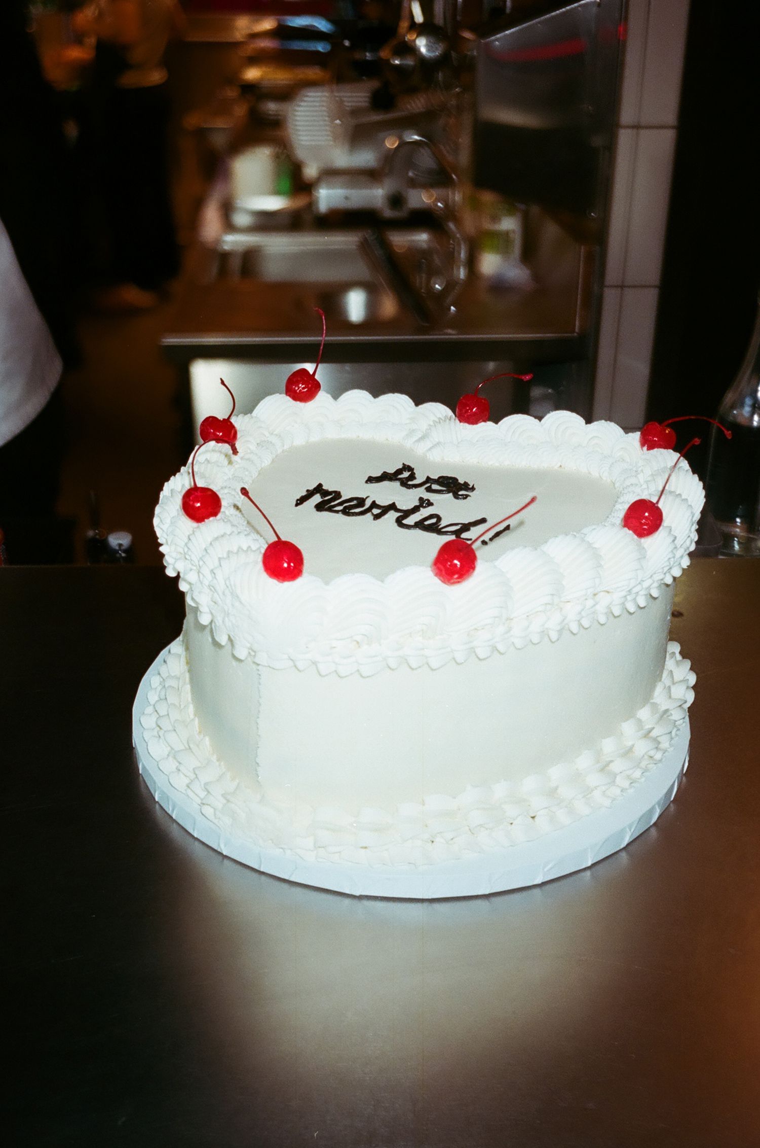 A white frosted cake decorated with red cherries and piped borders sits on a dark surface in a dimly lit setting.
