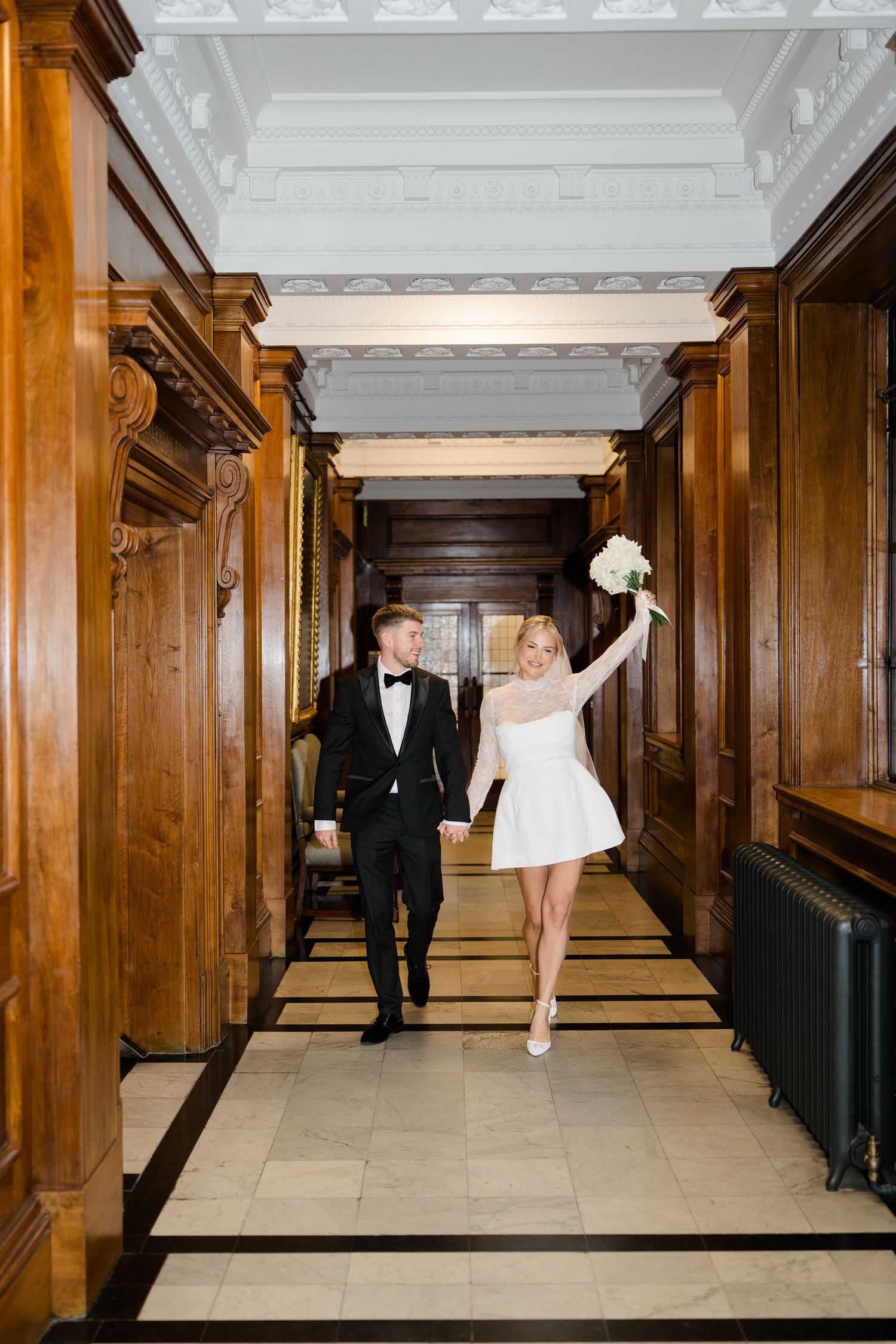 Wedding couple poses playfully in an elegant wood-paneled hallway with white ceiling.
