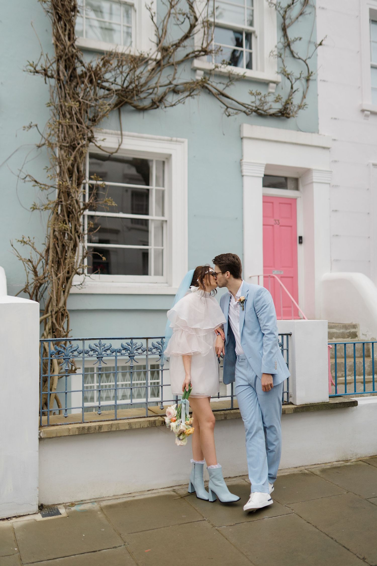 Couple kissing in front of a pastel blue townhouse with a pink door, bride in mini dress and groom in light blue suit.