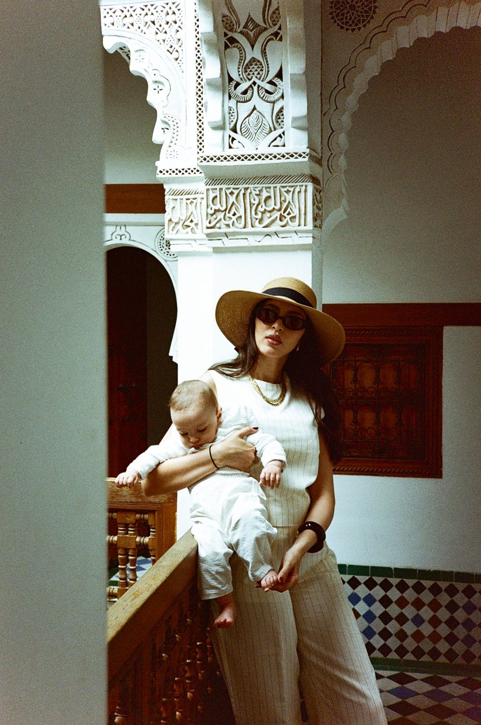 Woman in sun hat holds baby on ornate Moroccan riad balcony with intricate carved plasterwork.