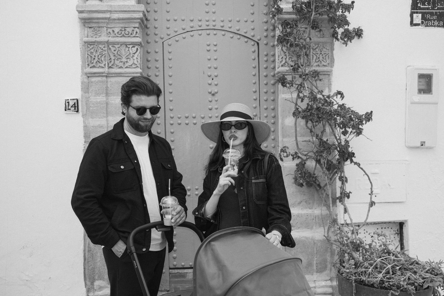 Black and white photo of a couple with a stroller walking near an ornate door in a European street.