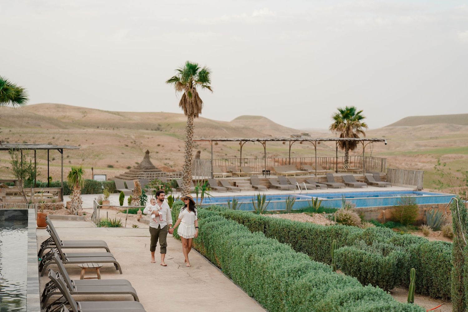 Couple walking along a desert resort pool surrounded by lush hedges and palm trees in an arid landscape.