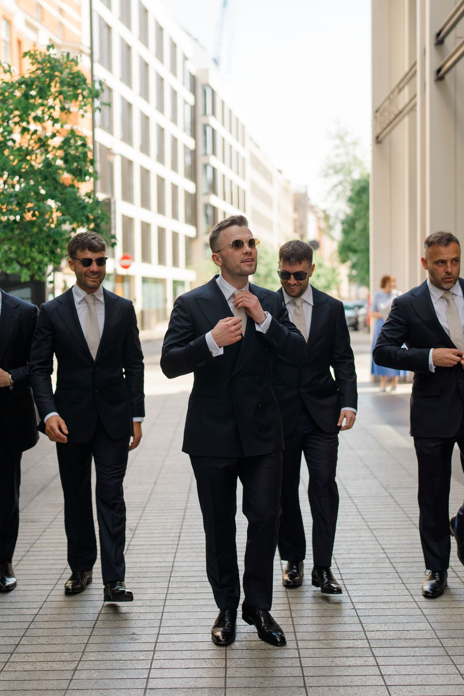 Groom and groomsmen in black suits and sunglasses walking confidently on a city street on wedding day.