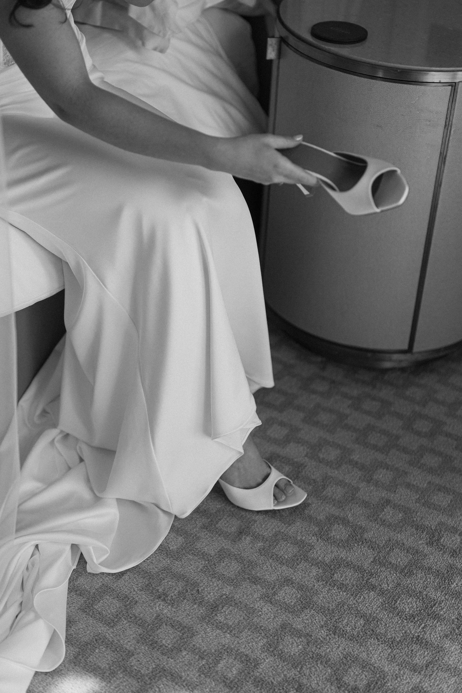 Black and white photo of a bride in a white gown putting on white heeled shoes while seated.