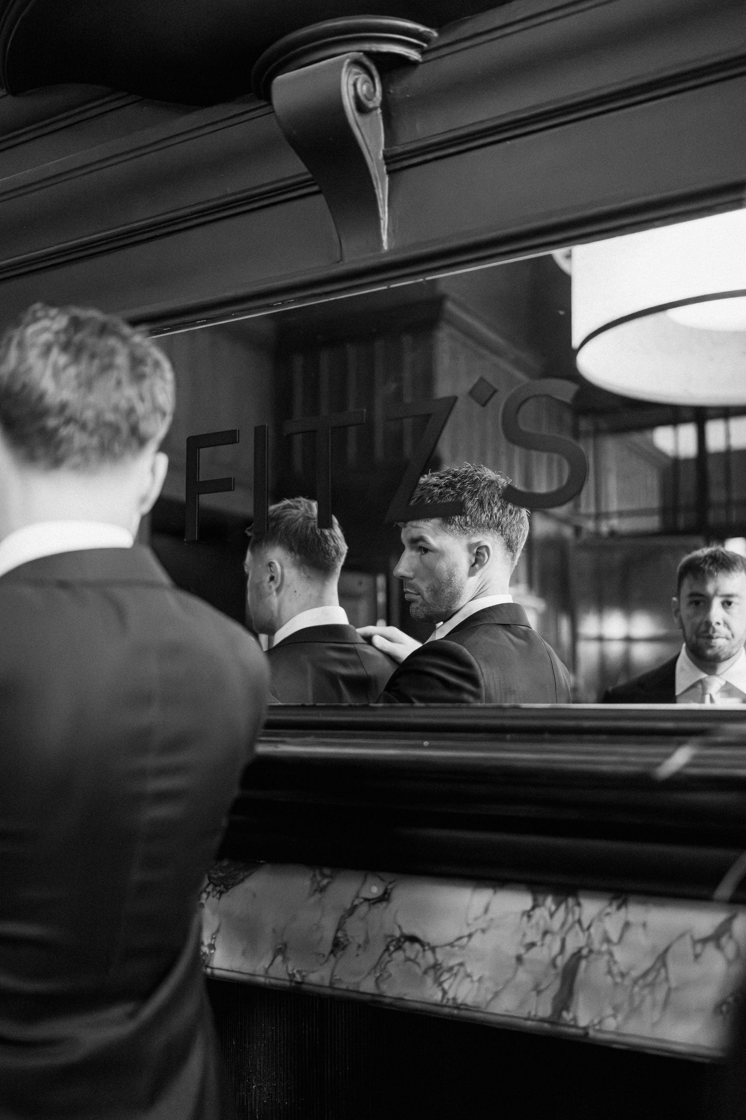 Groomsmen adjusting ties reflected in a bar mirror at a wedding venue.