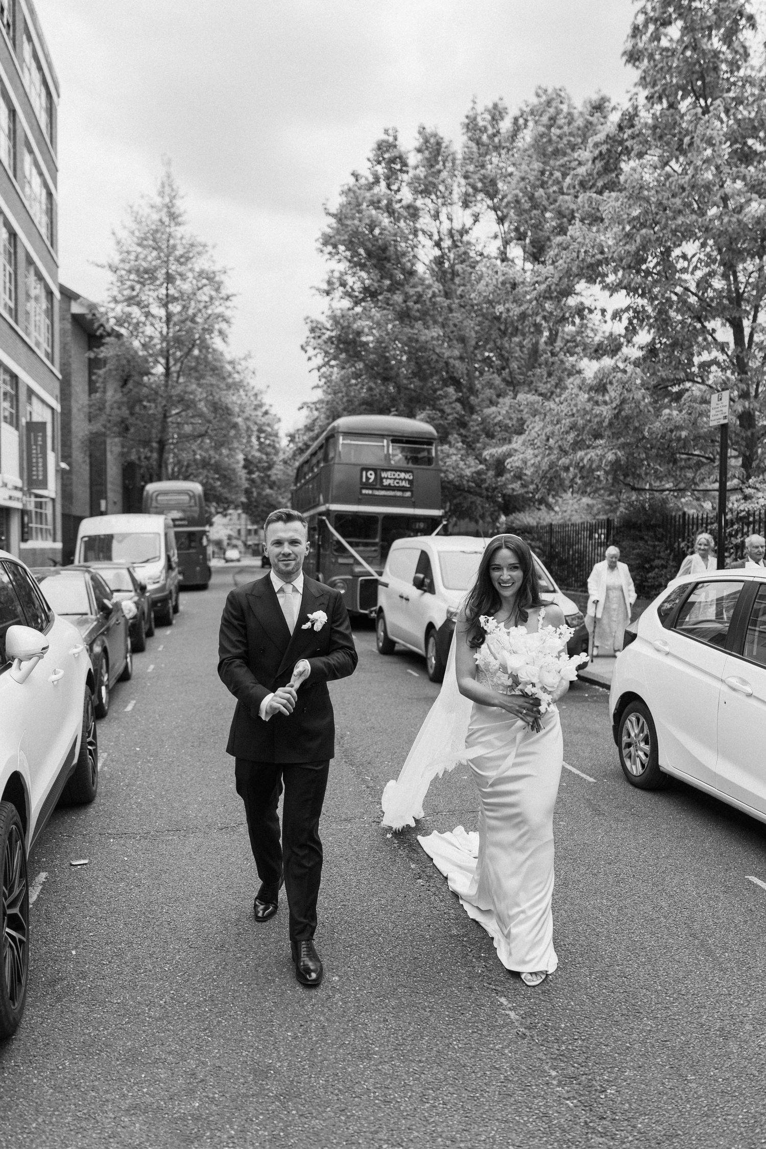 Black and white photo of bride and groom walking down a London street with a double-decker bus in the background.