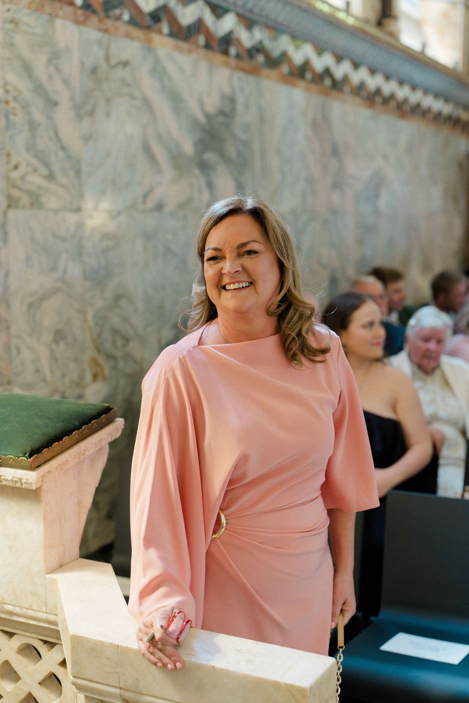 Smiling woman in elegant pink dress standing in a church during a wedding ceremony.