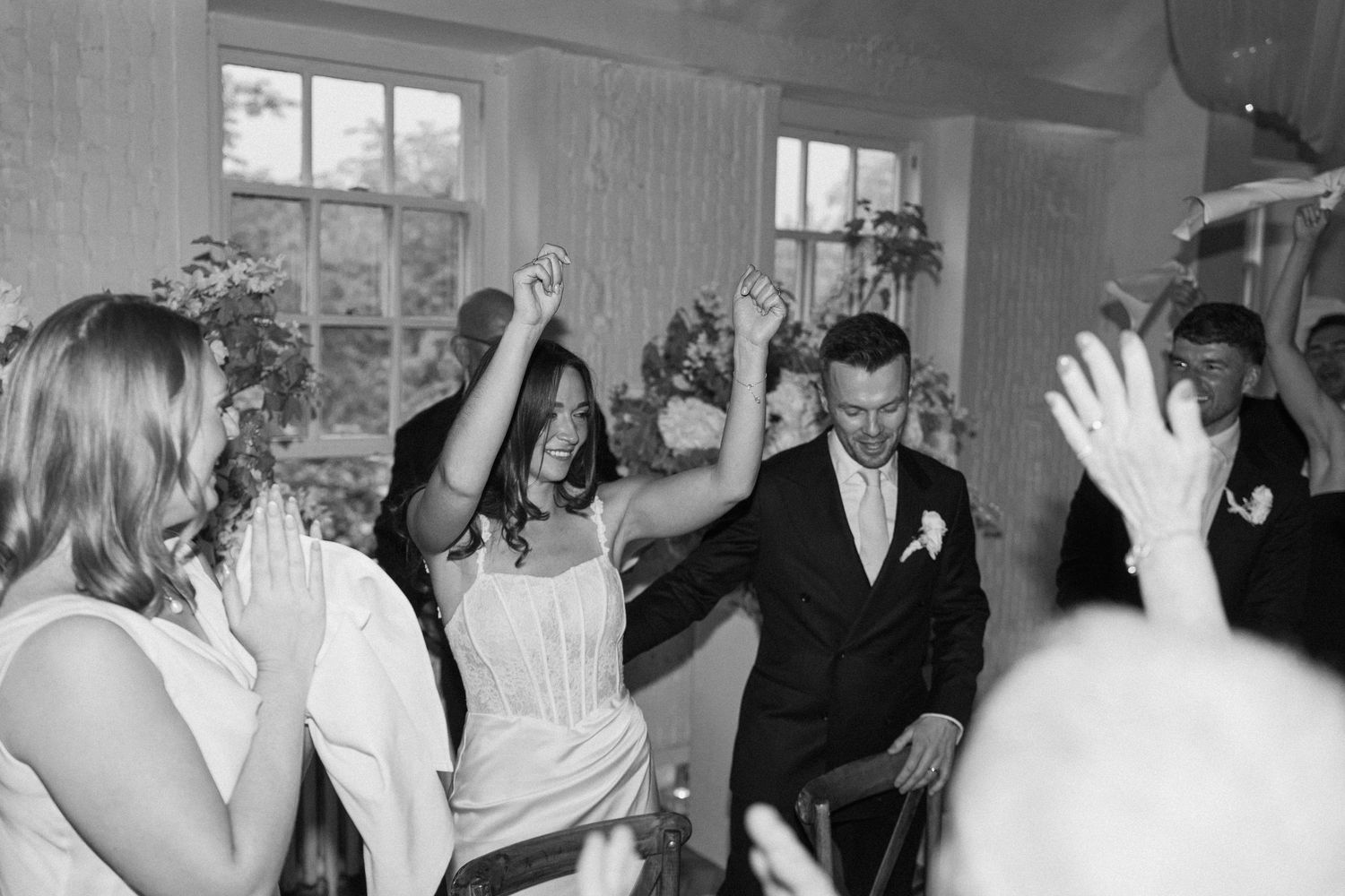 Joyful bride and groom dancing surrounded by cheering guests at lively wedding reception, black and white.