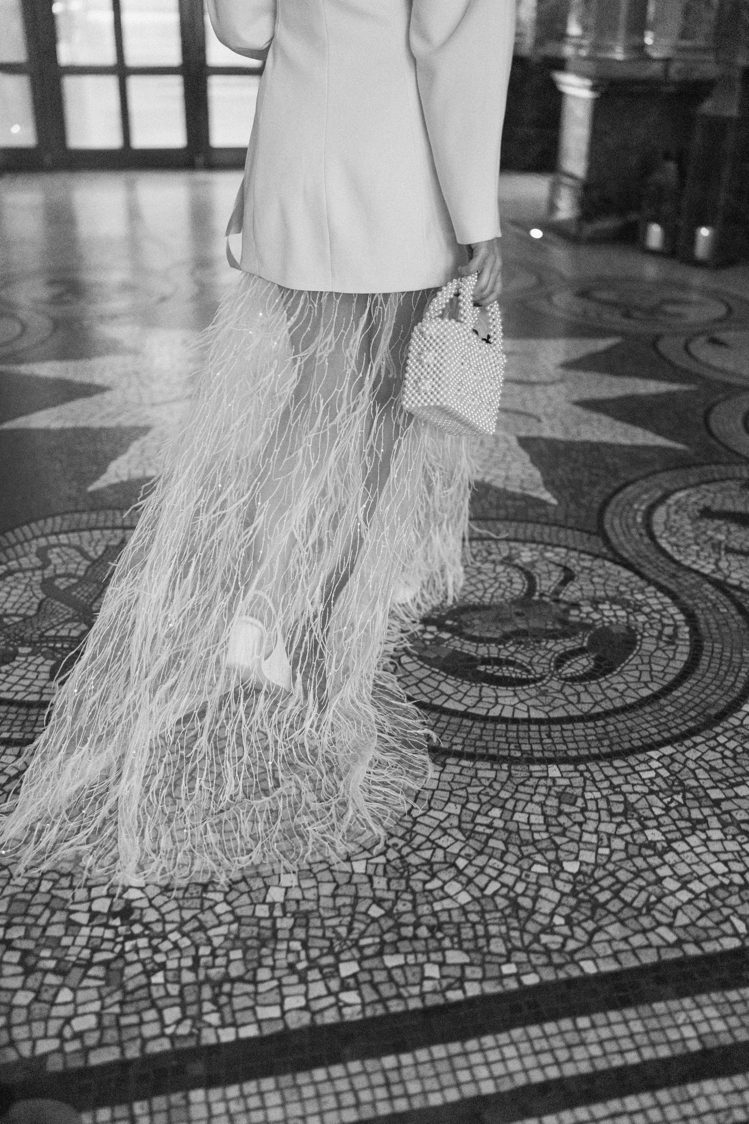 Black and white close-up of bride's feathered skirt and heels on mosaic tile floor.