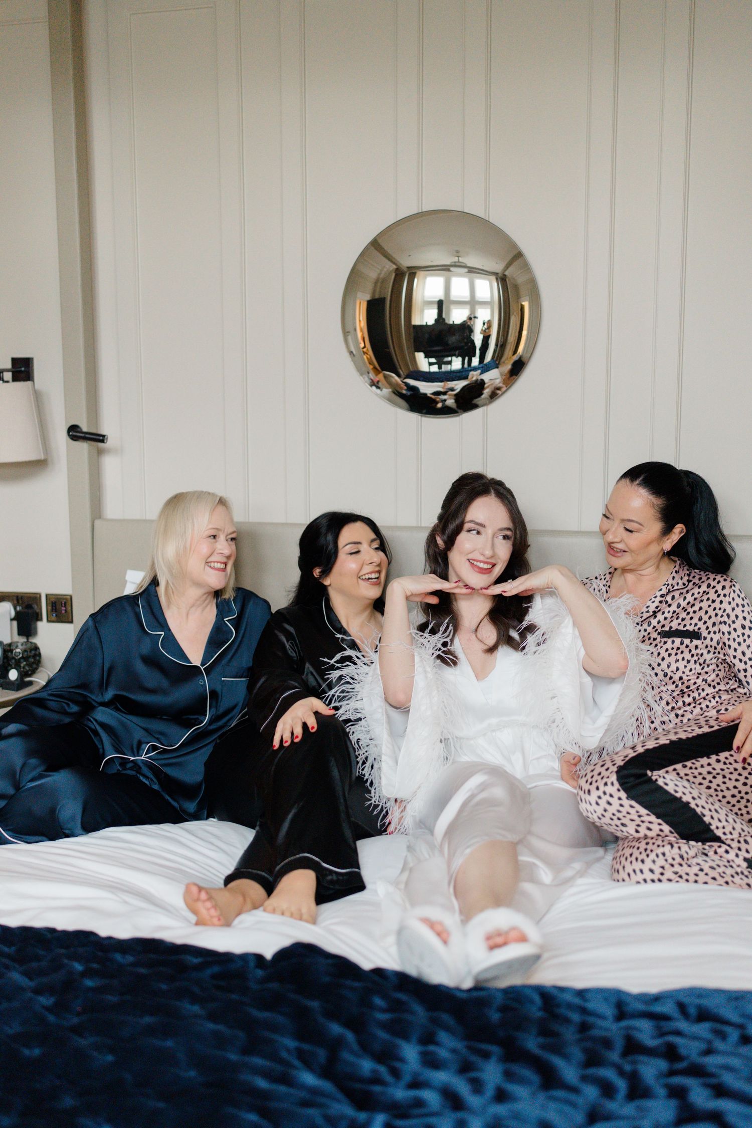 Bride in white feather robe sitting on bed with bridesmaids in satin pajamas laughing.