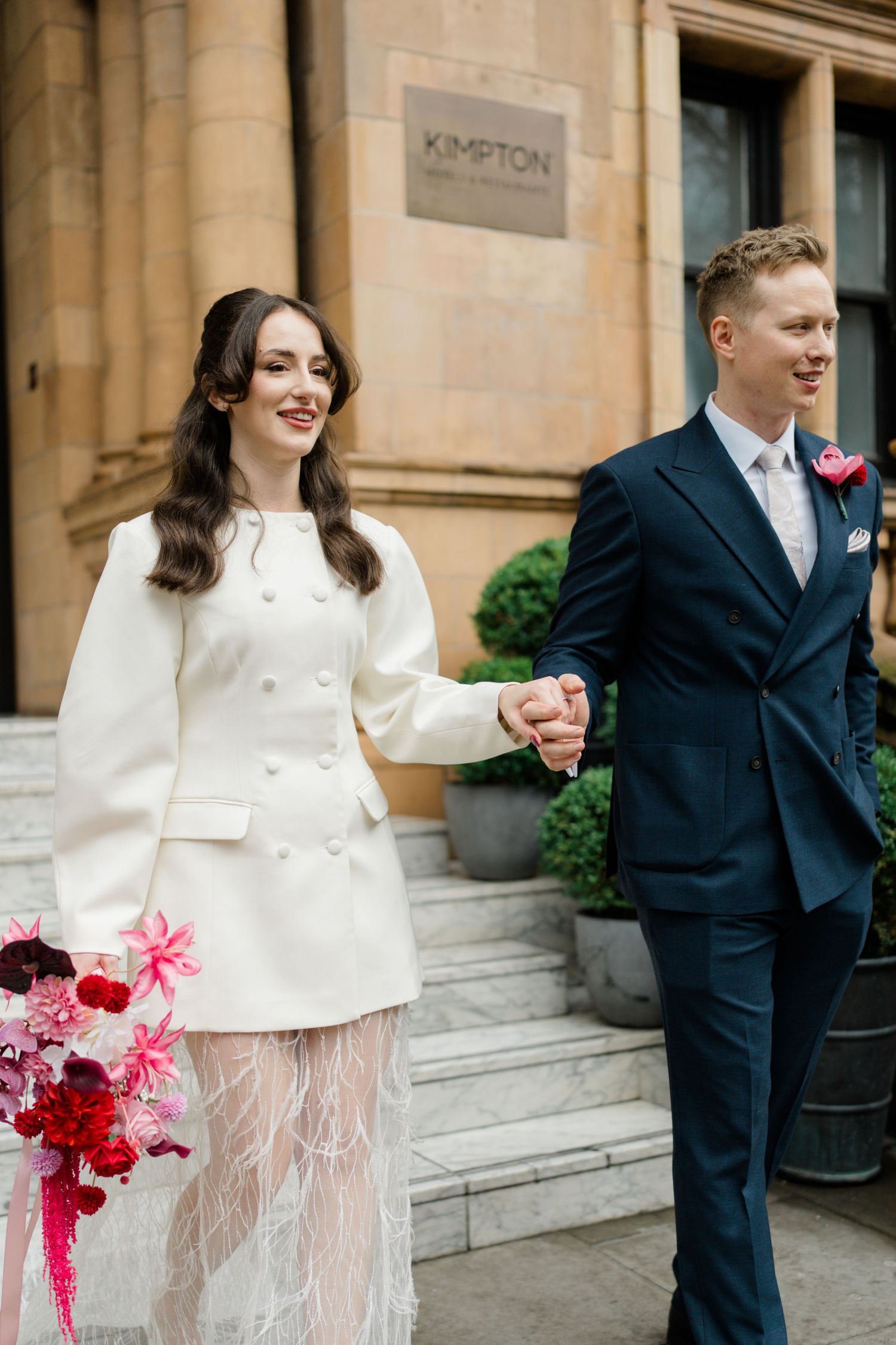 Happy newlyweds holding hands outside Kimpton Hotel, bride in white coat dress carrying pink floral bouquet.