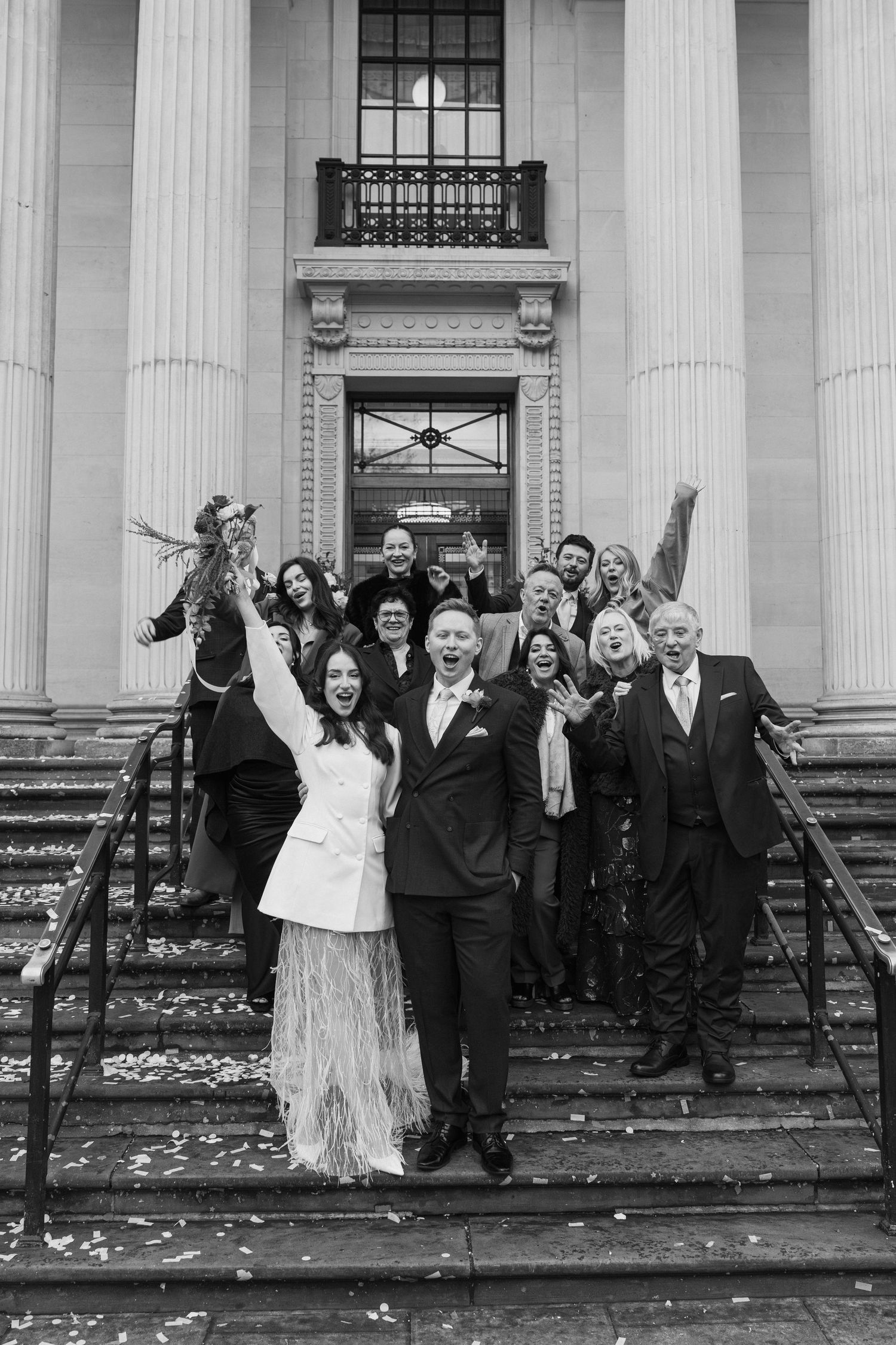 Black and white photo of joyful wedding party celebrating with confetti on grand building steps.