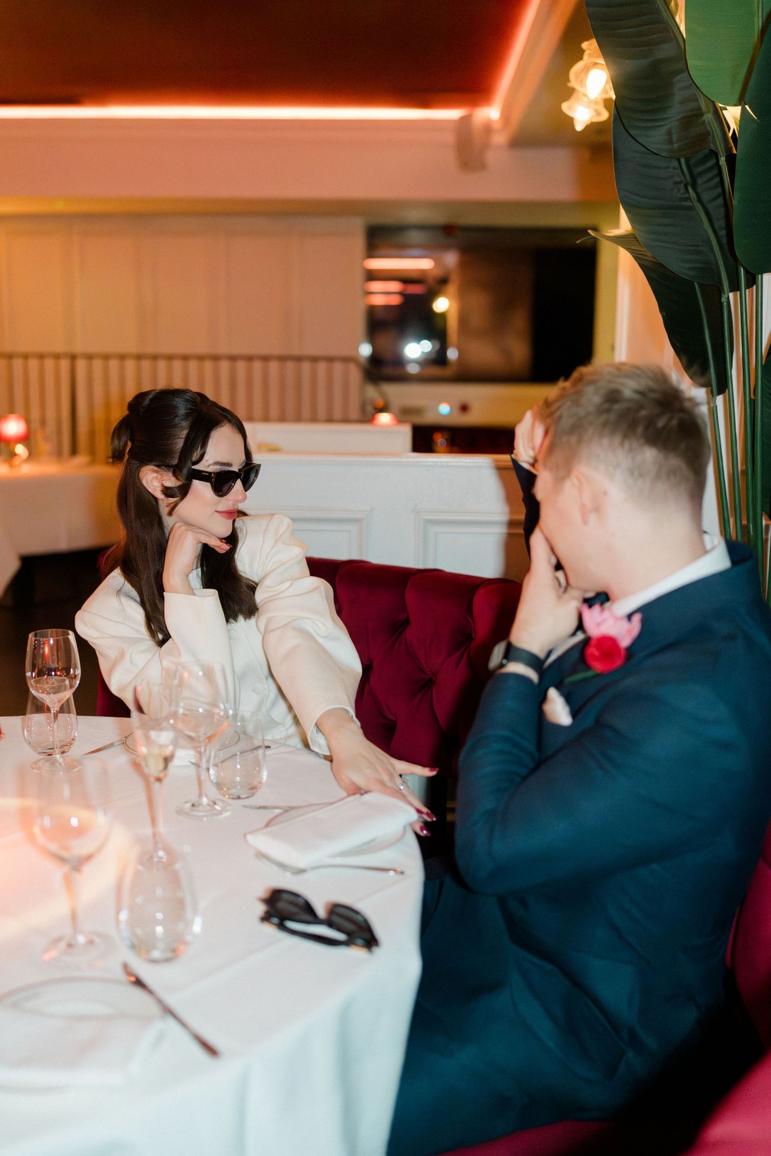 Stylish bride in sunglasses and white blazer seated with groom at elegant restaurant table.