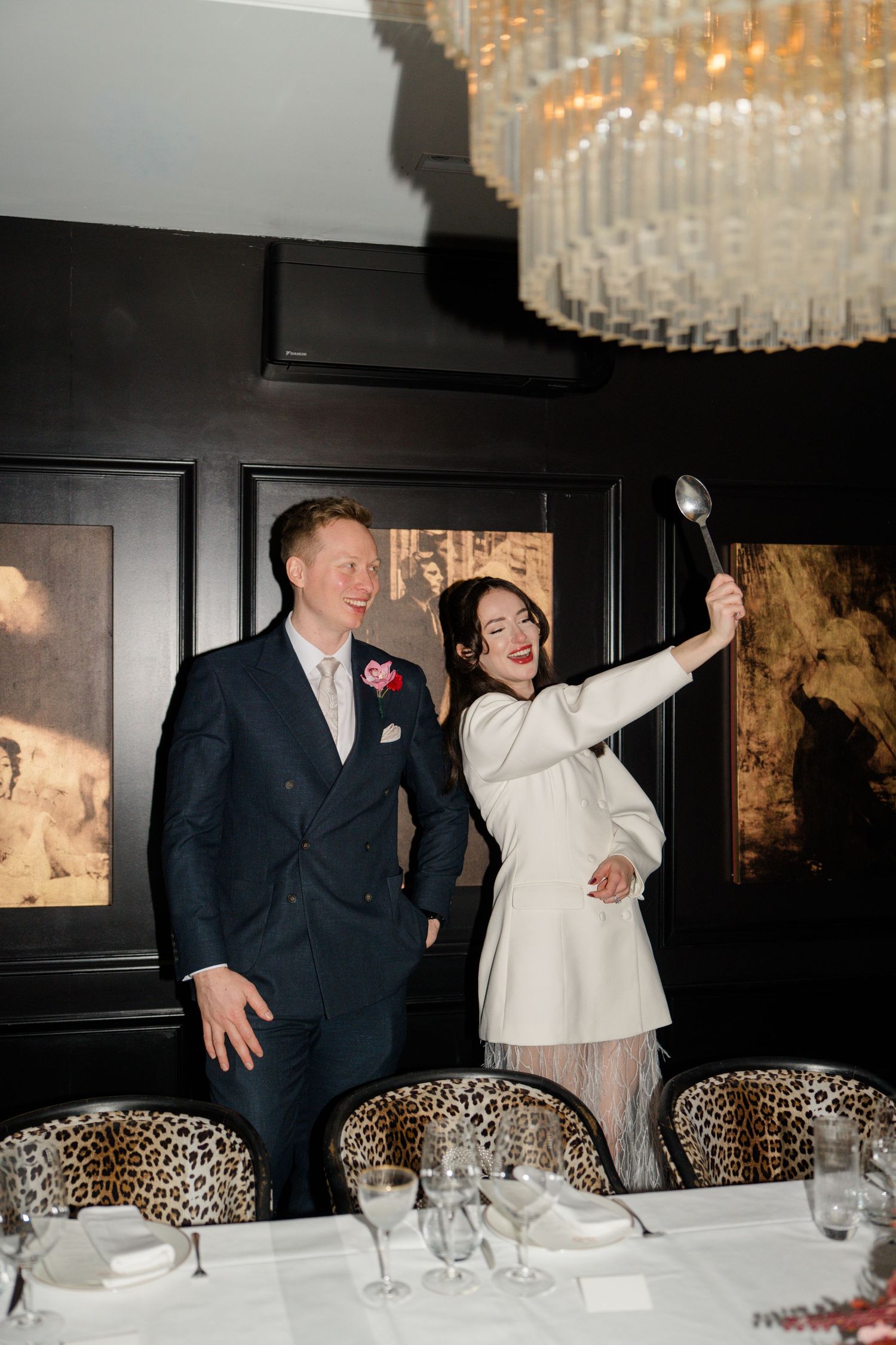 Laughing bride in white mini dress playfully raises a spoon beside her groom in a dark elegant dining room.