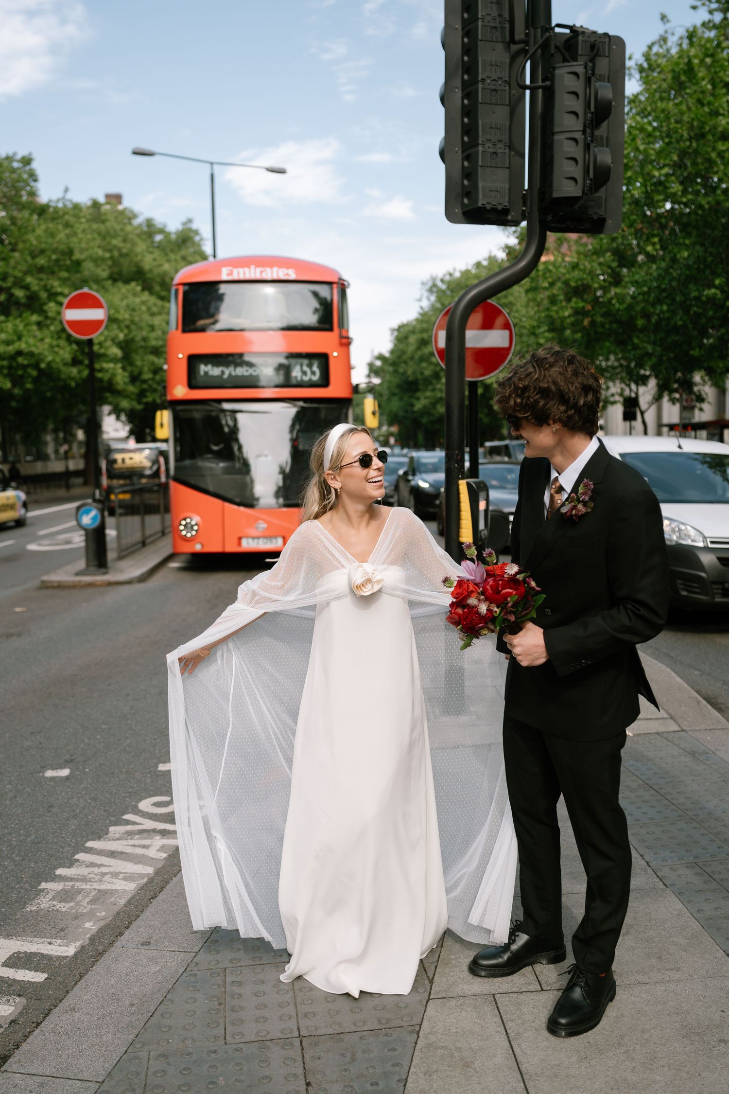 A couple poses playfully on a London street with a red bus in the background.