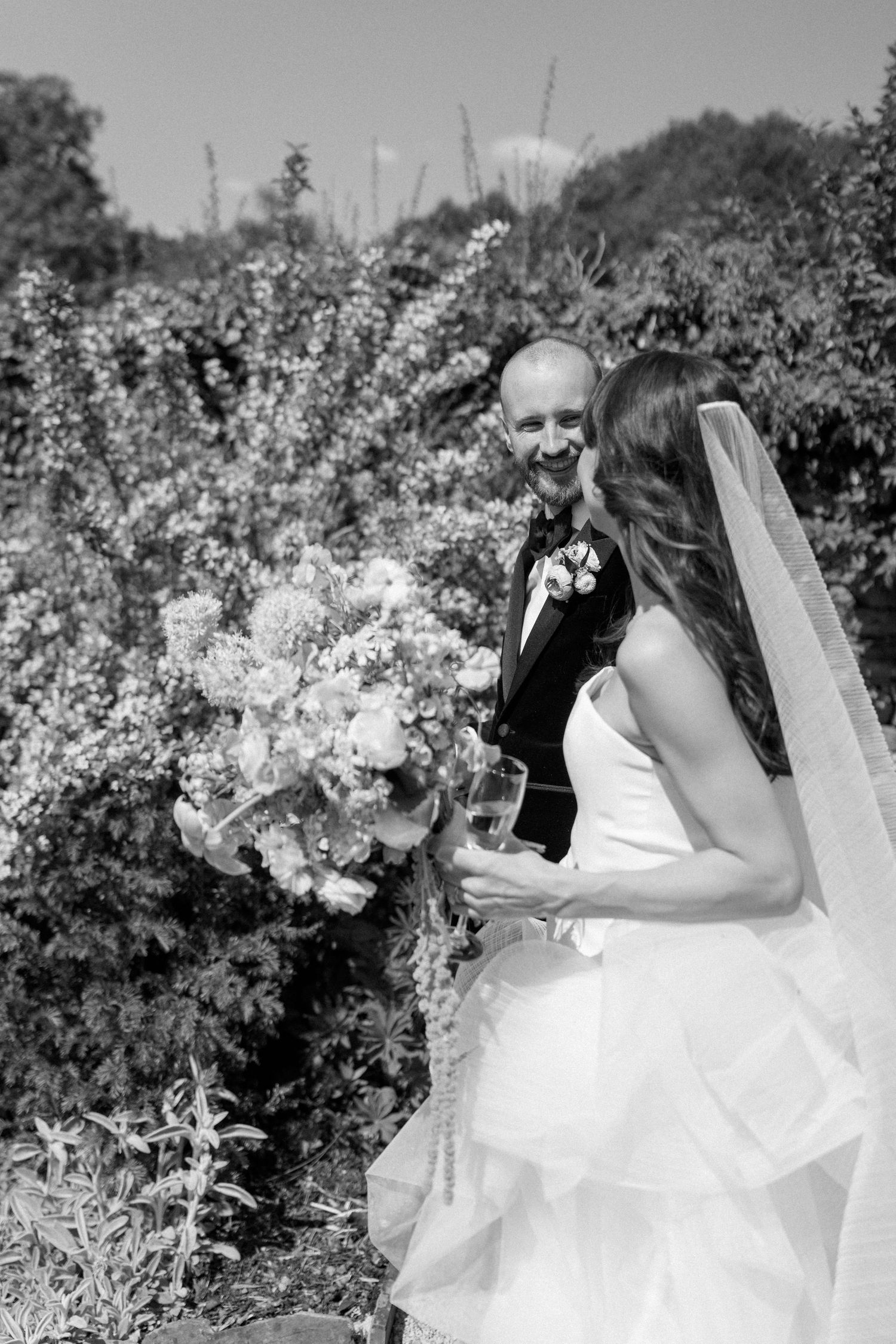 A black and white wedding photo captures a couple walking through a garden.