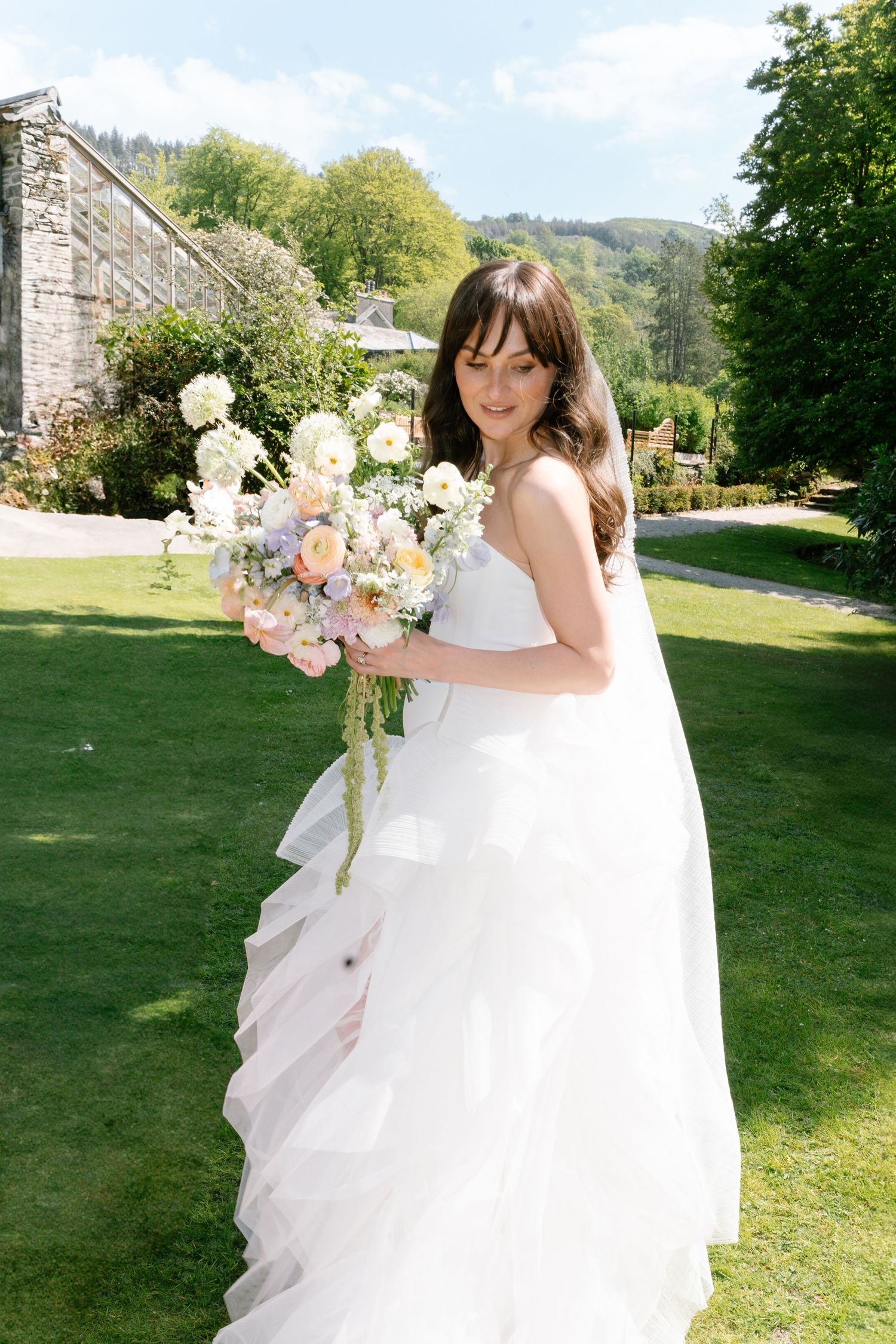 A bride in a flowing white gown poses with her bouquet on a sunny lawn.